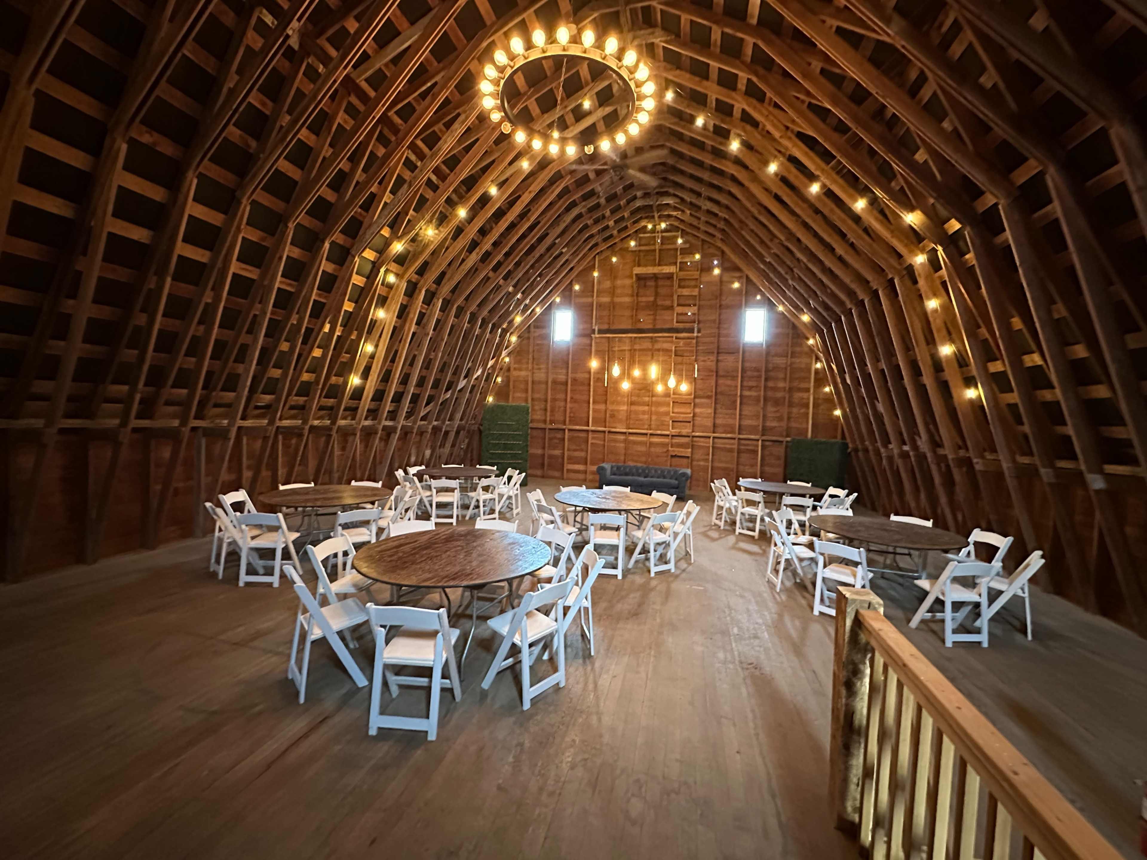The interior of a barn features circular tables with white chairs arranged around them, under a wooden ceiling adorned with decorative lights.