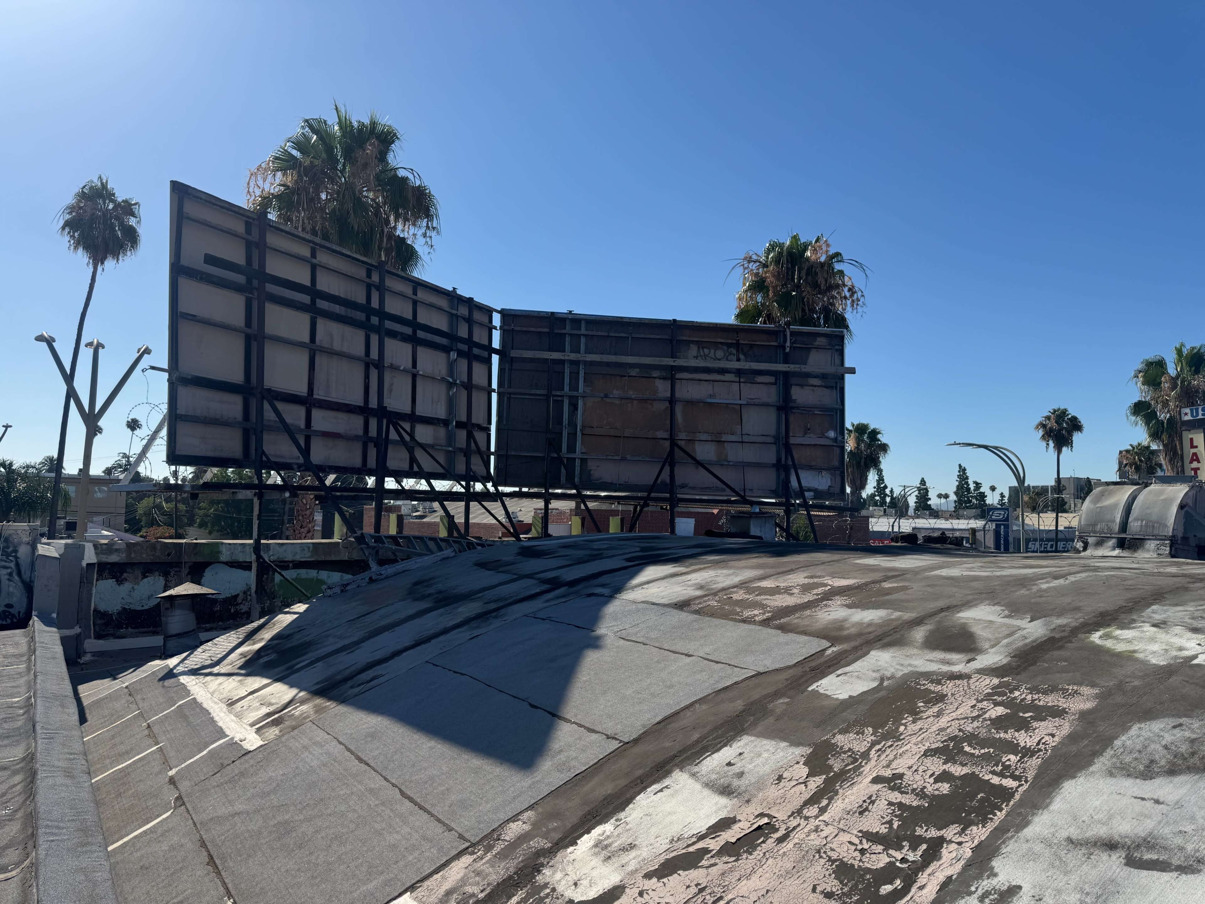 The image shows a rooftop with two large, empty billboards against a clear blue sky, surrounded by palm trees.