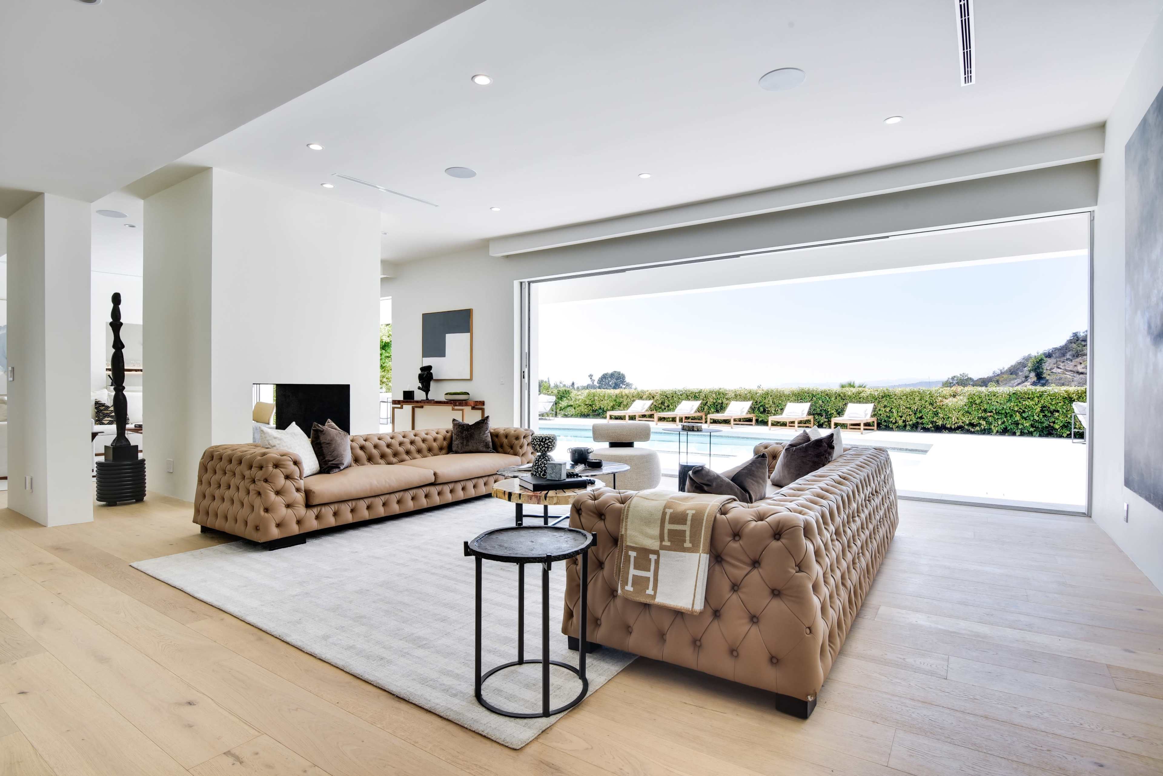 A spacious living room features two tufted leather sofas, a coffee table, and large floor-to-ceiling glass doors opening to an outdoor terrace with a view of the landscape.