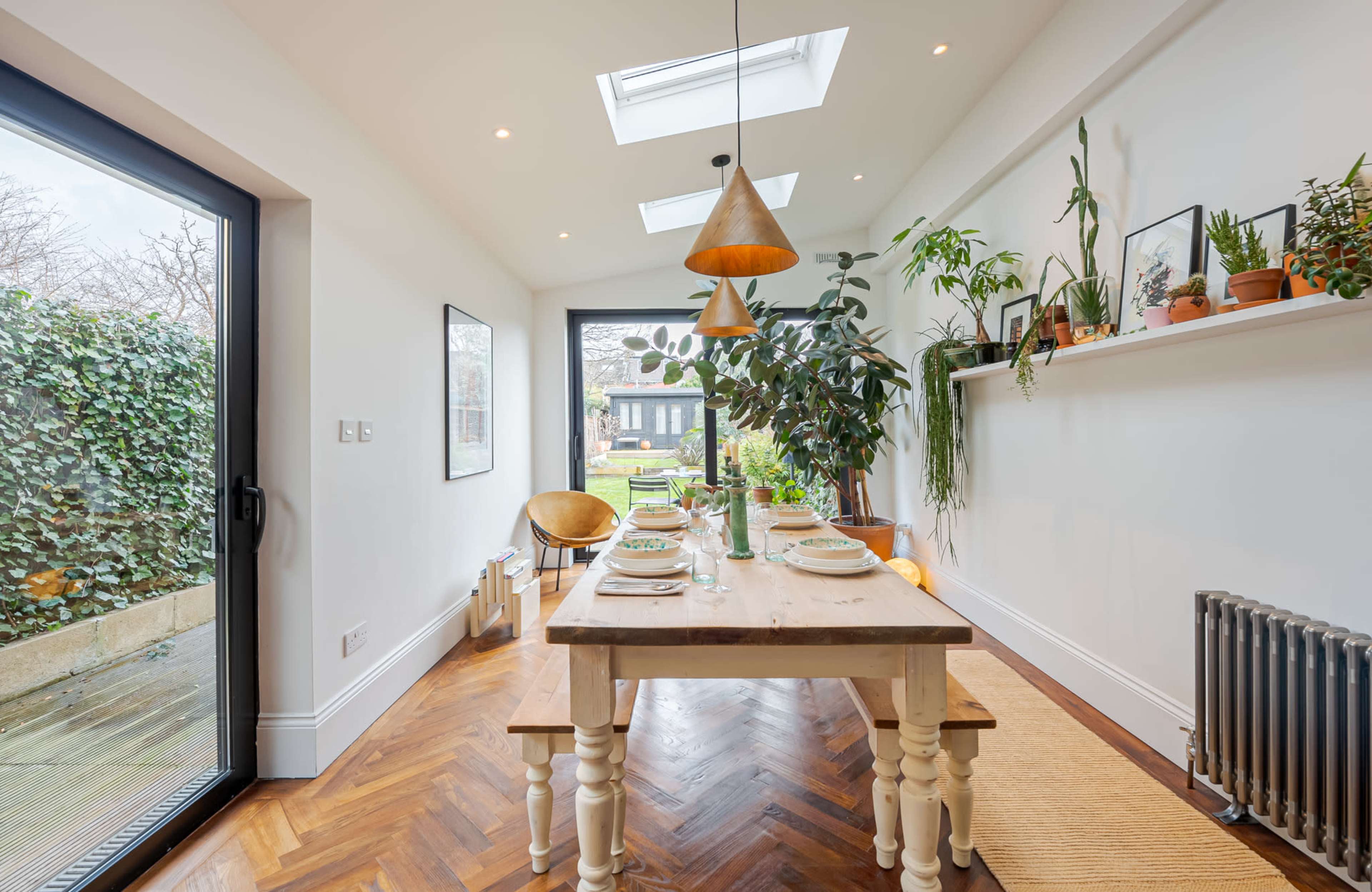 A dining area features a wooden table set for a meal, surrounded by plants and large windows that overlook a garden.