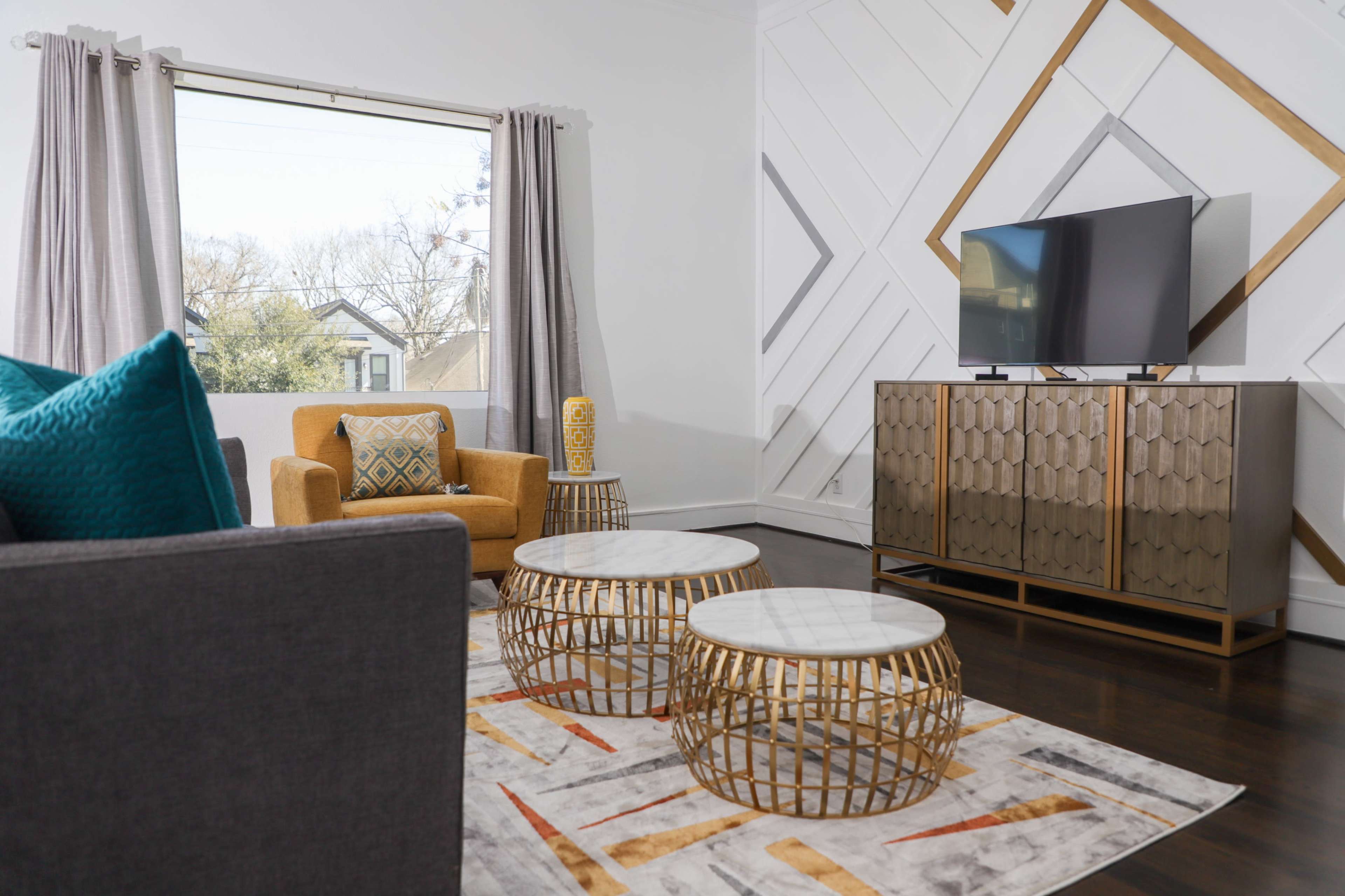 A modern living room with a gray sofa, an orange armchair, a television on a stylish cabinet, and decorative tables on a patterned rug.