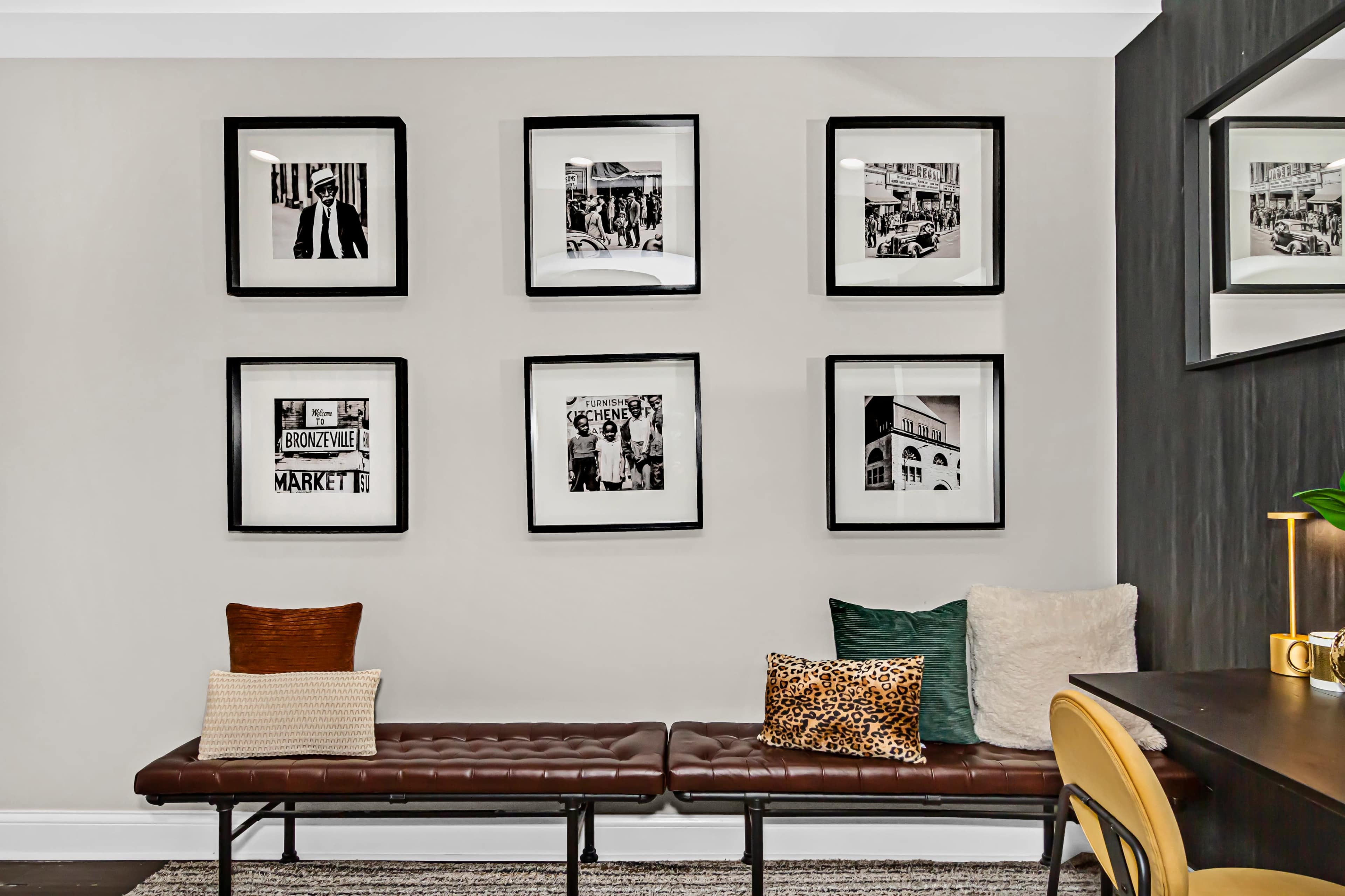The scene features a wall display with eight framed black-and-white photographs above a brown bench, surrounded by decorative pillows, in a modern room.