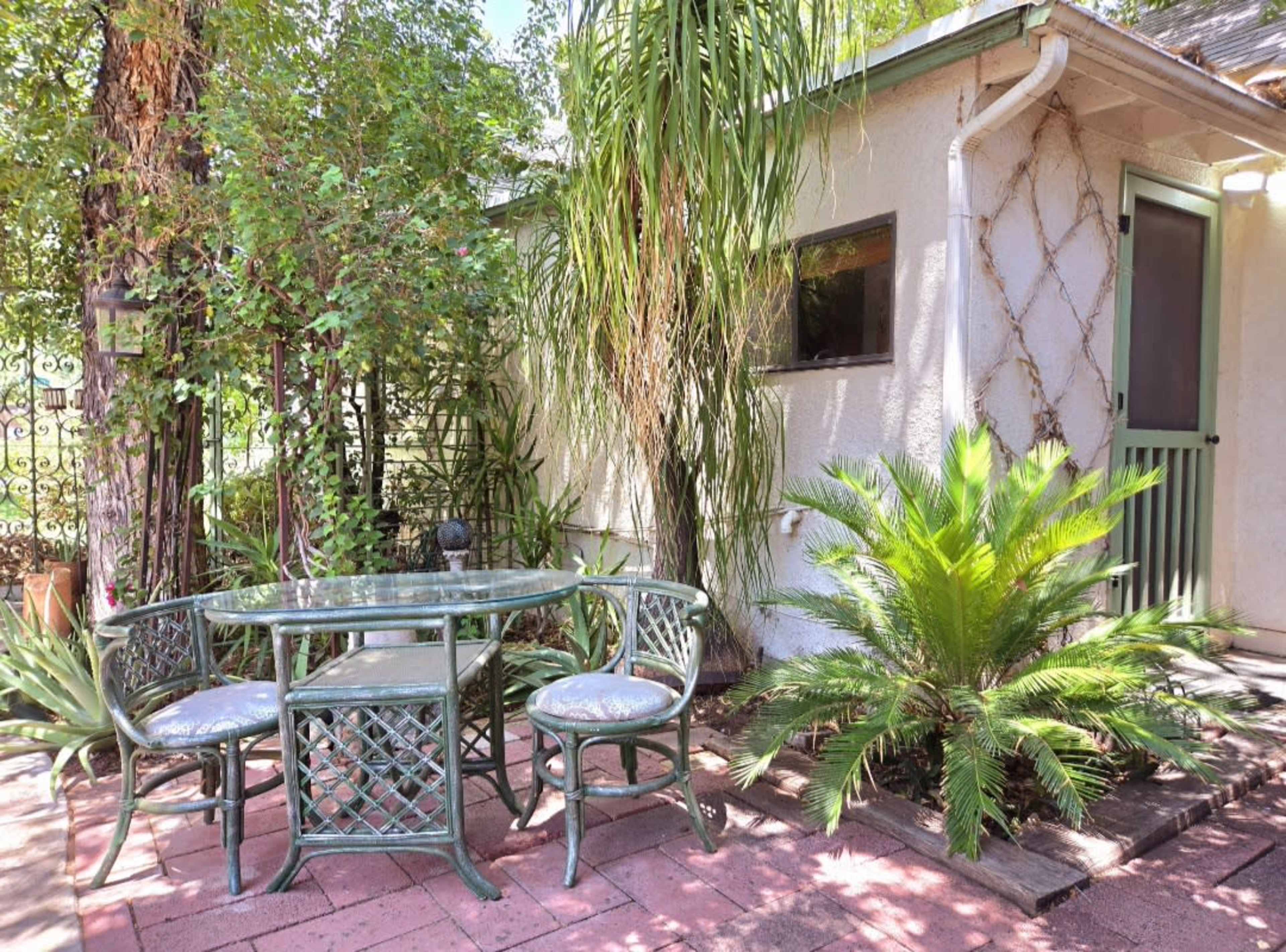 A small outdoor seating area features a glass table surrounded by two wrought-iron chairs, with potted plants and greenery framing the space.