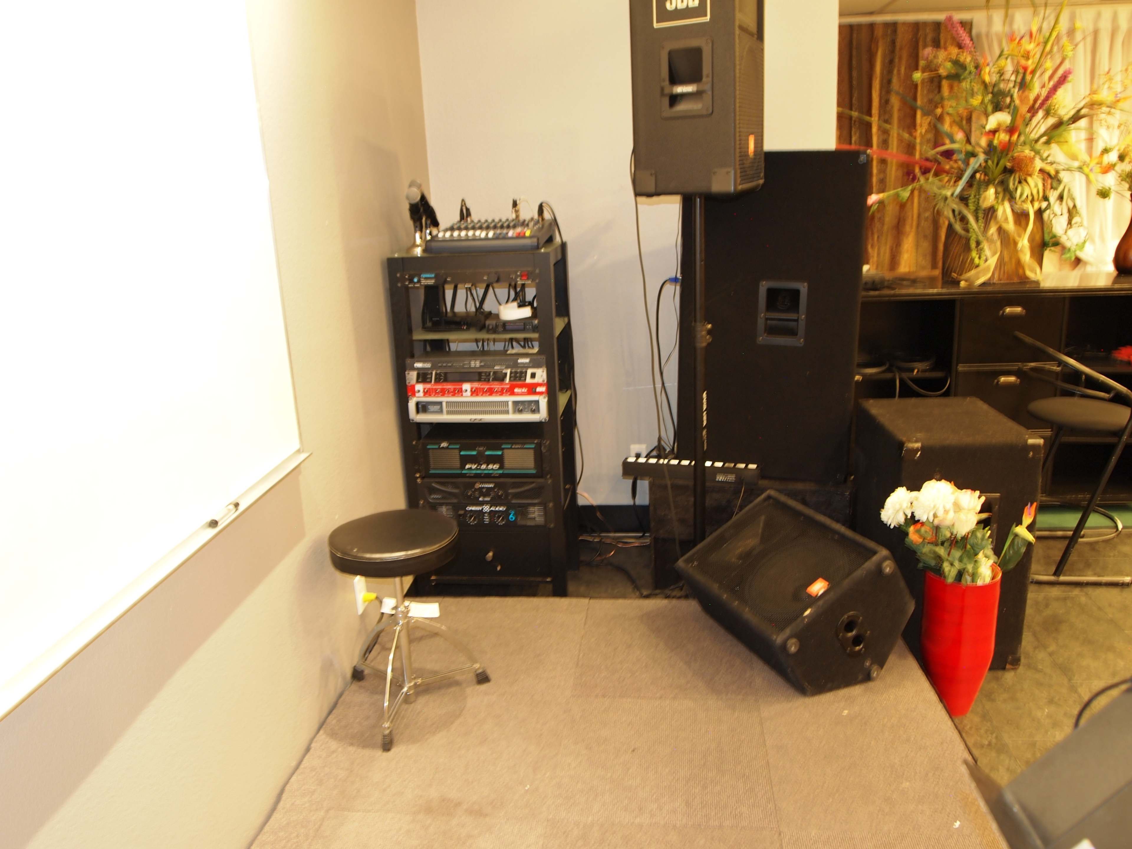 A music equipment setup in a corner of a room, featuring speakers, a rack of audio gear, a black stool, and a flower arrangement in a red vase.