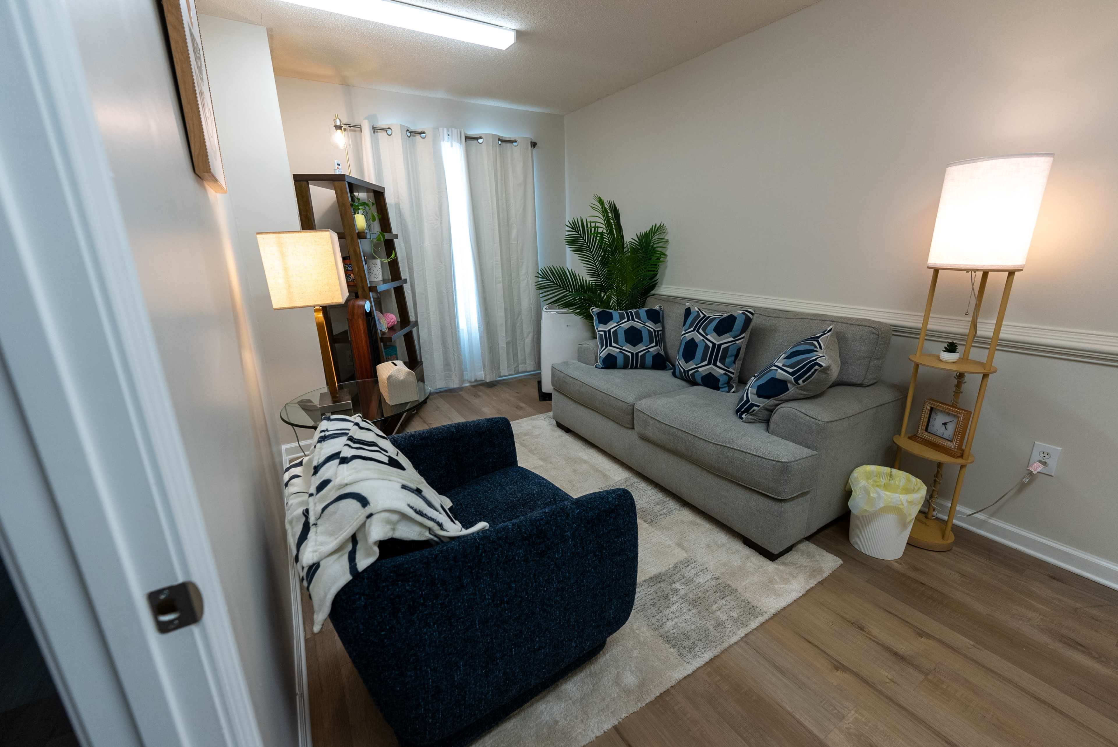 The image shows a cozy living room with a gray sofa, a blue accent chair, a coffee table, and a floor lamp, along with a plant and a bookshelf against the wall.