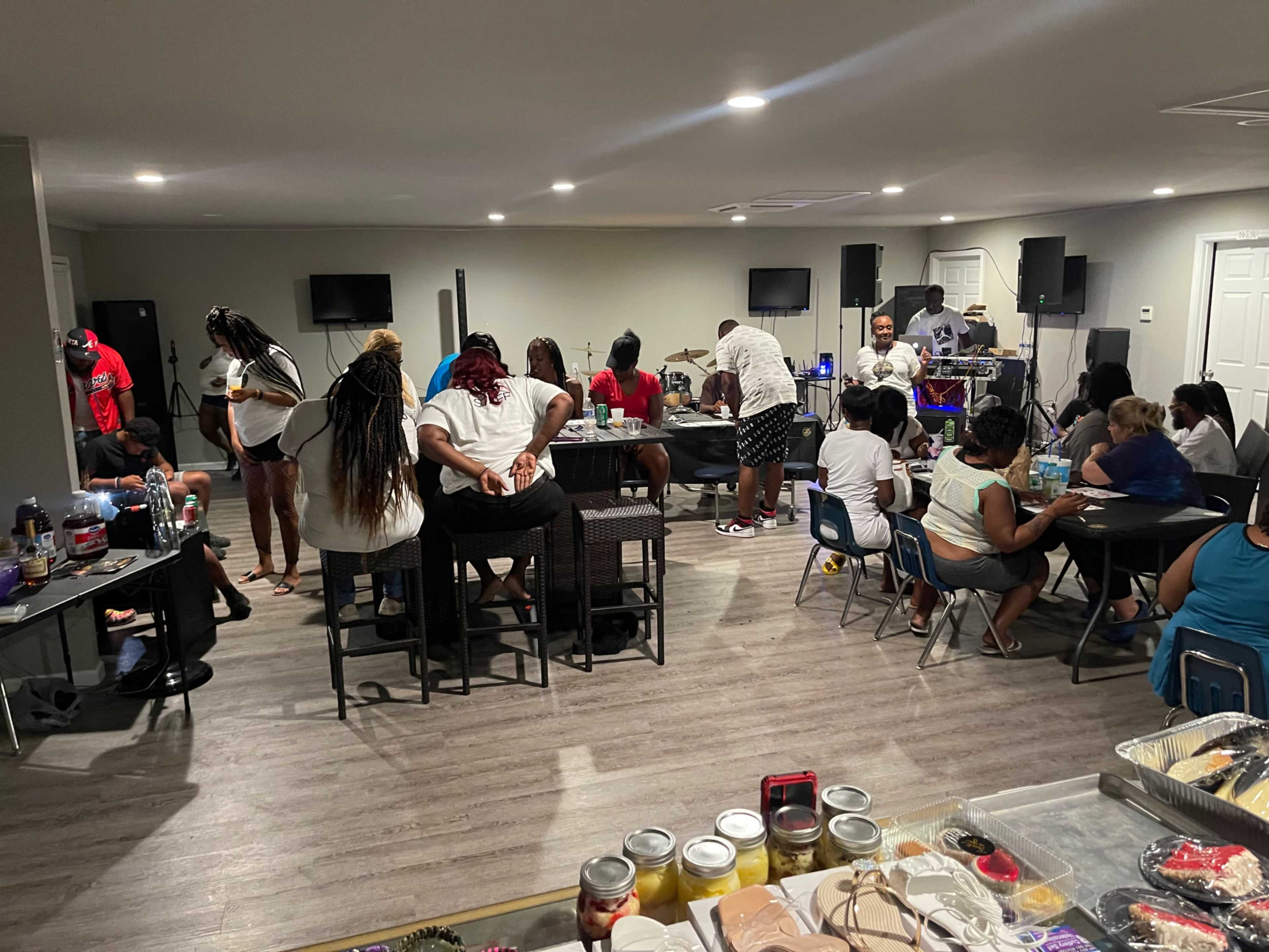 A group of people are enjoying a social gathering in a spacious room with tables and chairs, while others are performing music in the background.