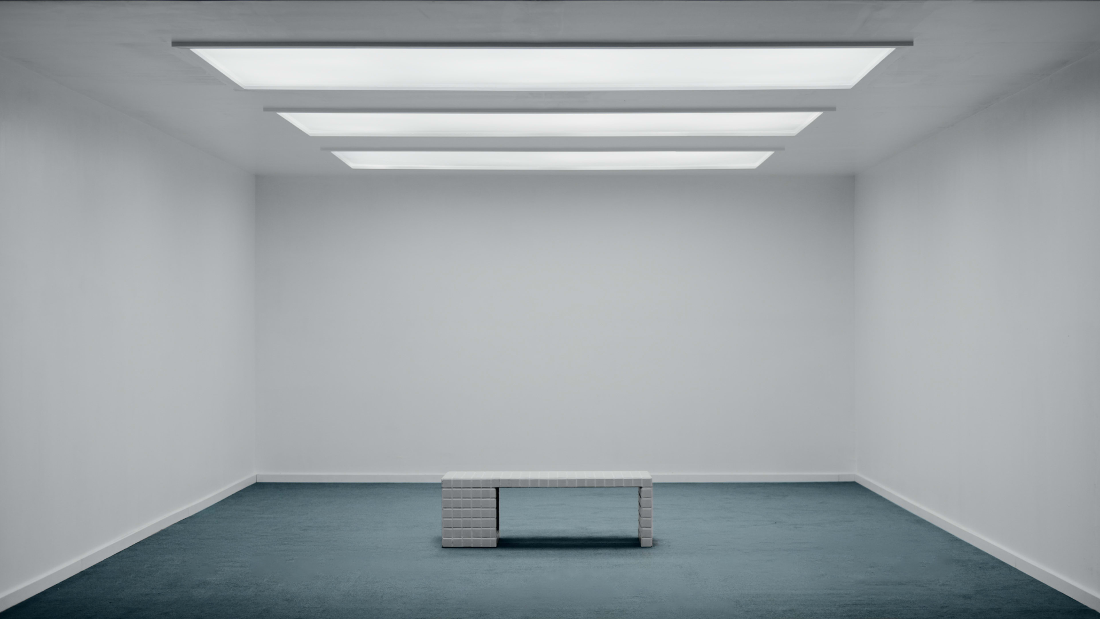 A stark, minimalist room features a white bench in the center and large, rectangular skylights above, illuminating the space.