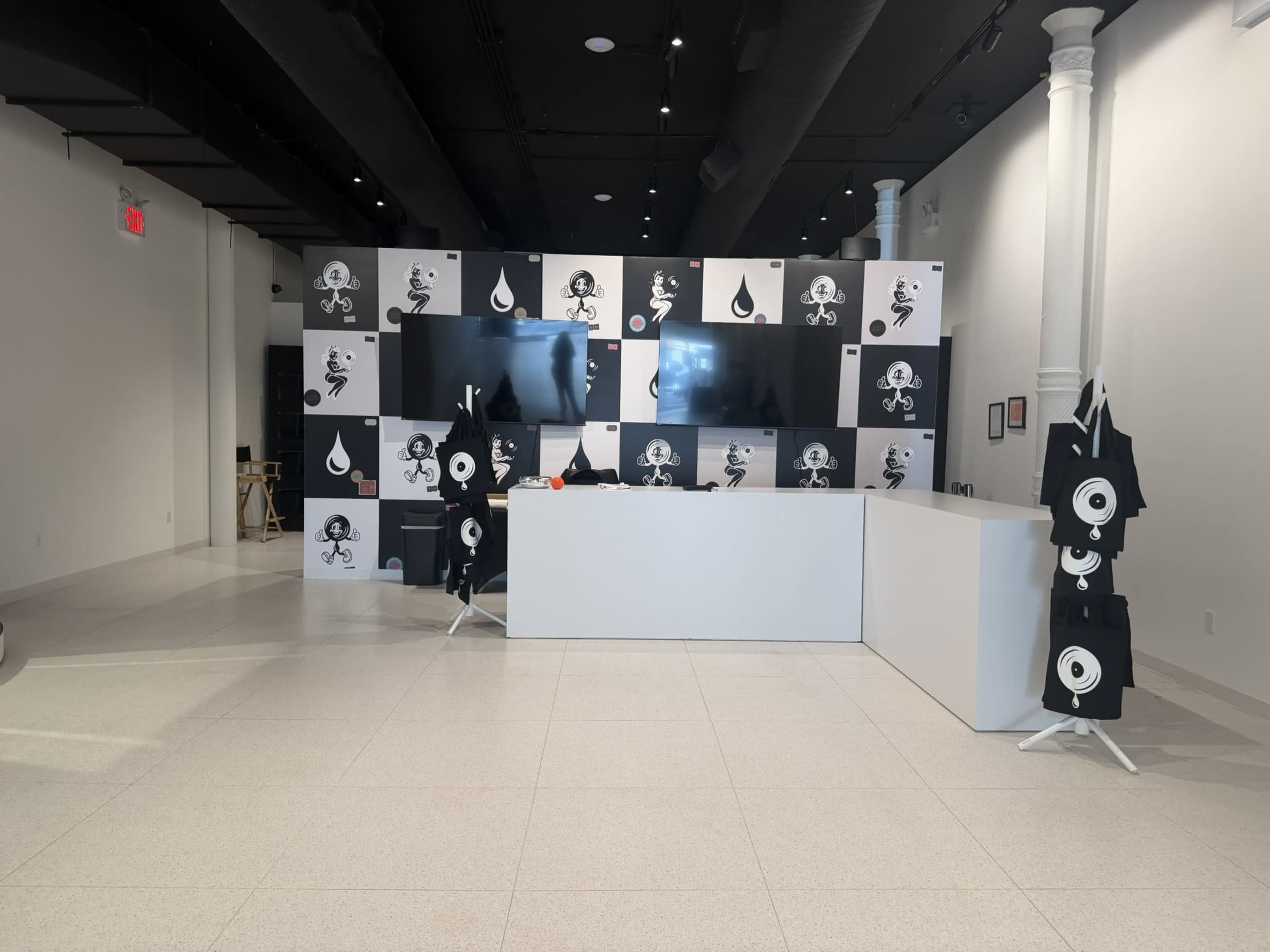 The image shows a modern exhibition space with a reception desk and a wall covered in black and white artwork featuring various illustrated designs.