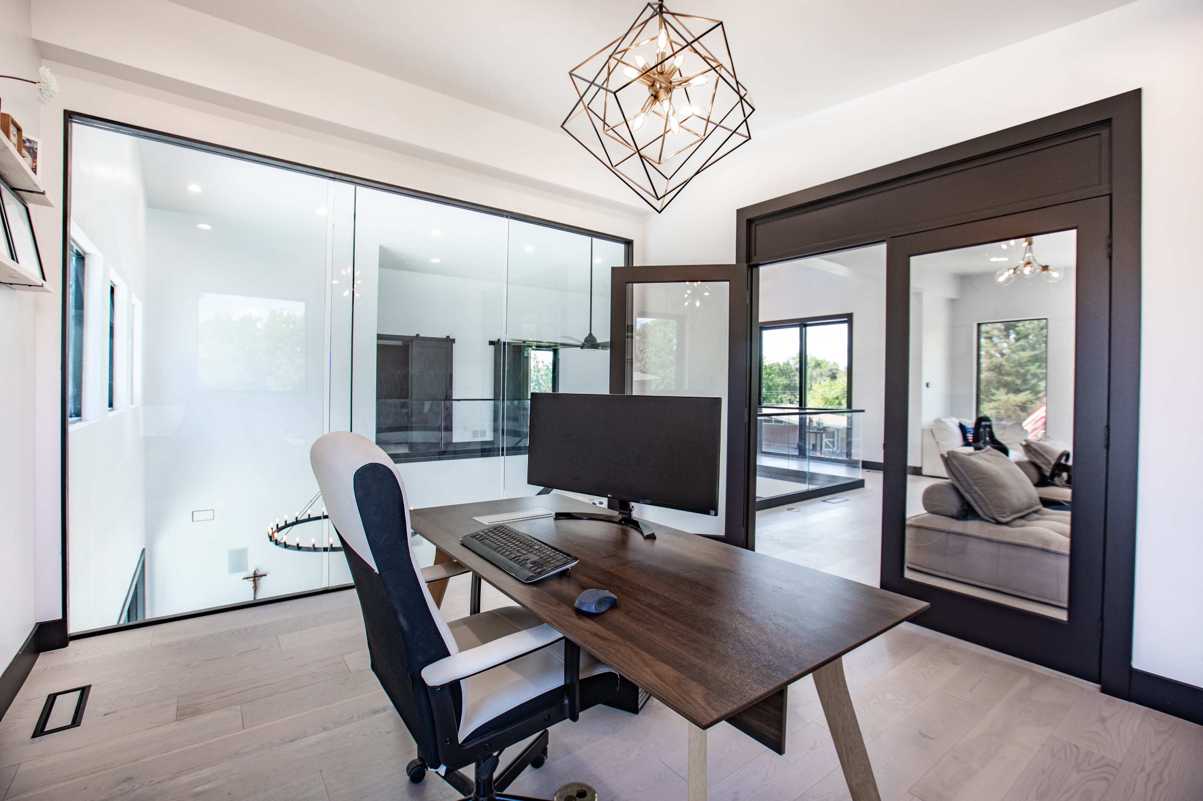 A modern home office features a wooden desk with a monitor and an ergonomic chair, enclosed by glass walls showcasing a bright interior.