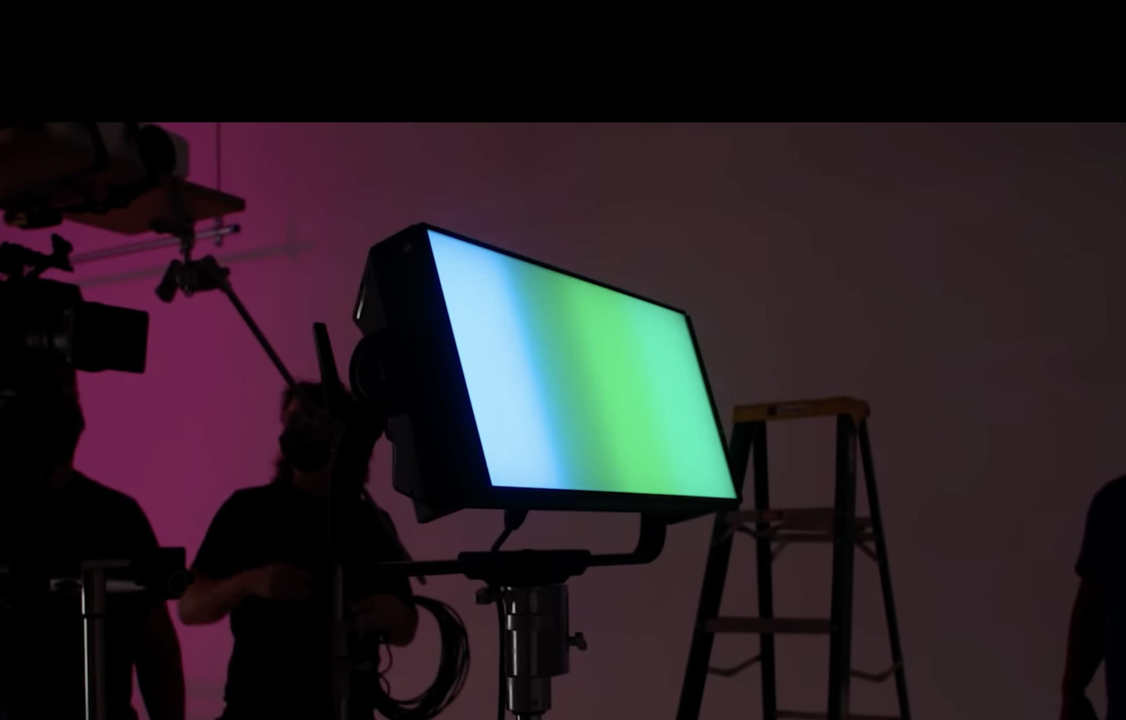 A color-changing light panel is positioned on a stand in a studio, with crew members and a ladder visible in the background.