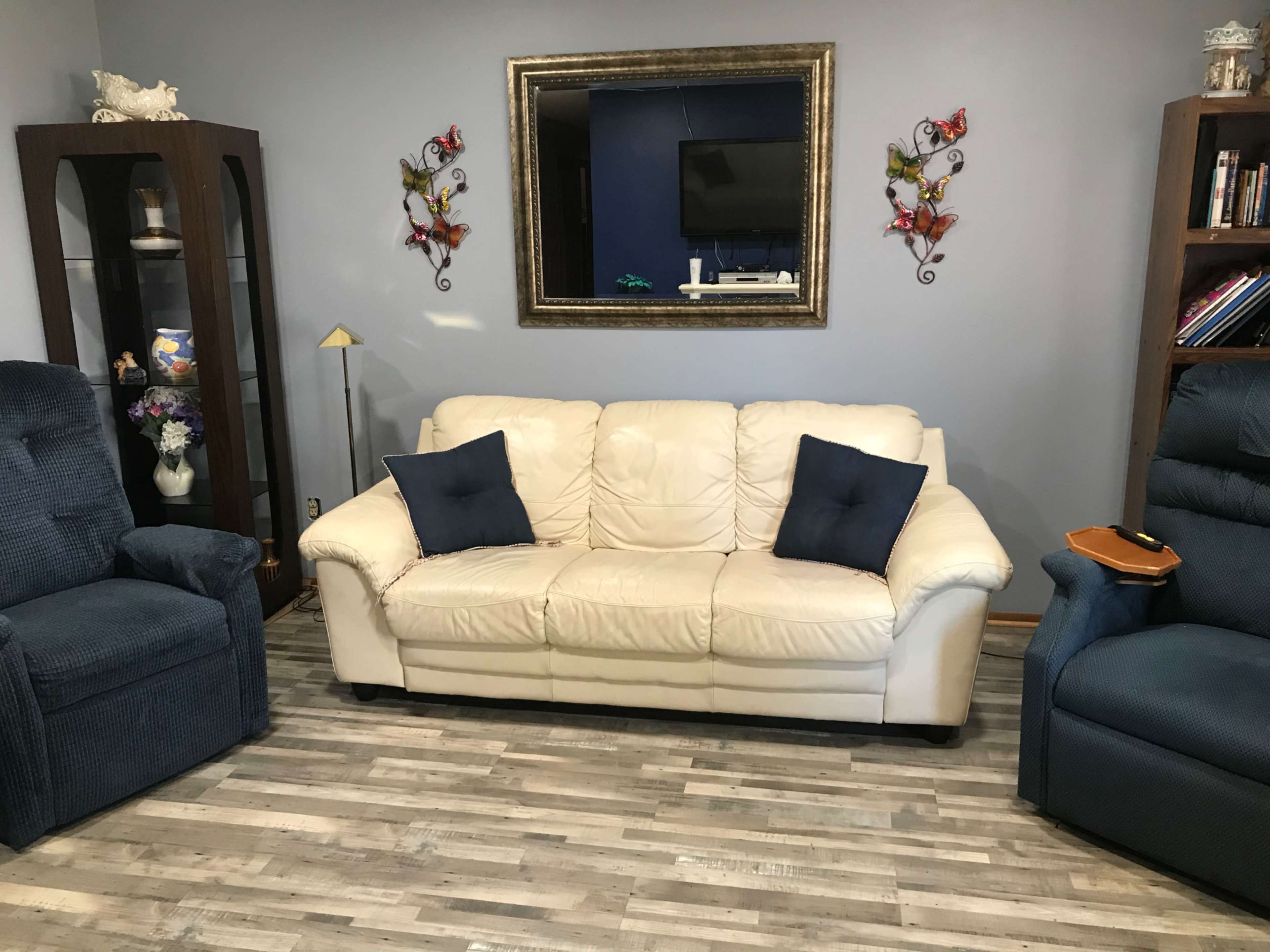 A living room with a white leather couch, two upholstered armchairs, a large mirror on the wall, and decorative items including butterflies and a lamp.