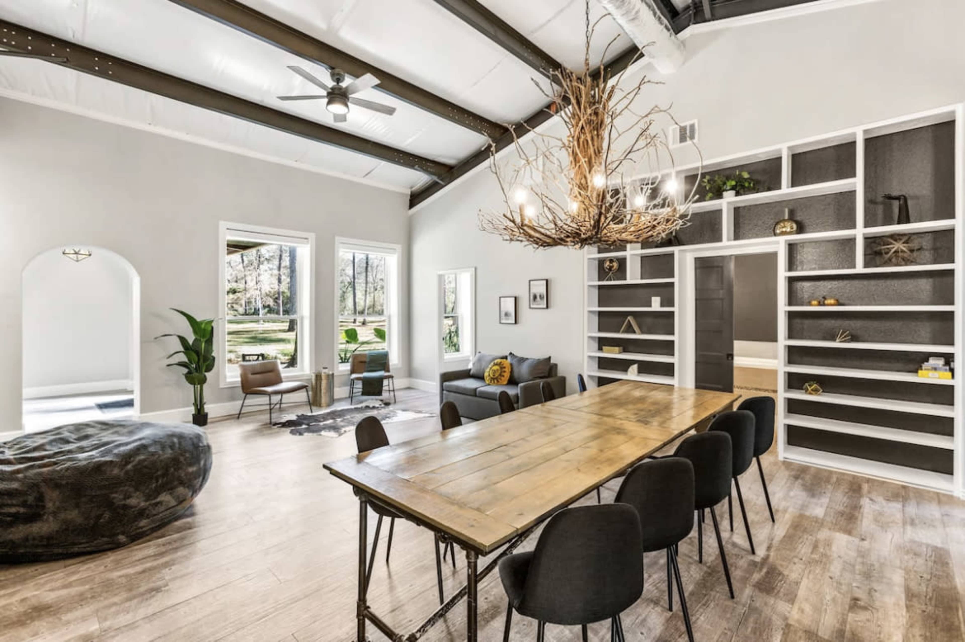 A spacious living area features a large wooden dining table, black chairs, and a comfortable seating area with large windows letting in natural light.