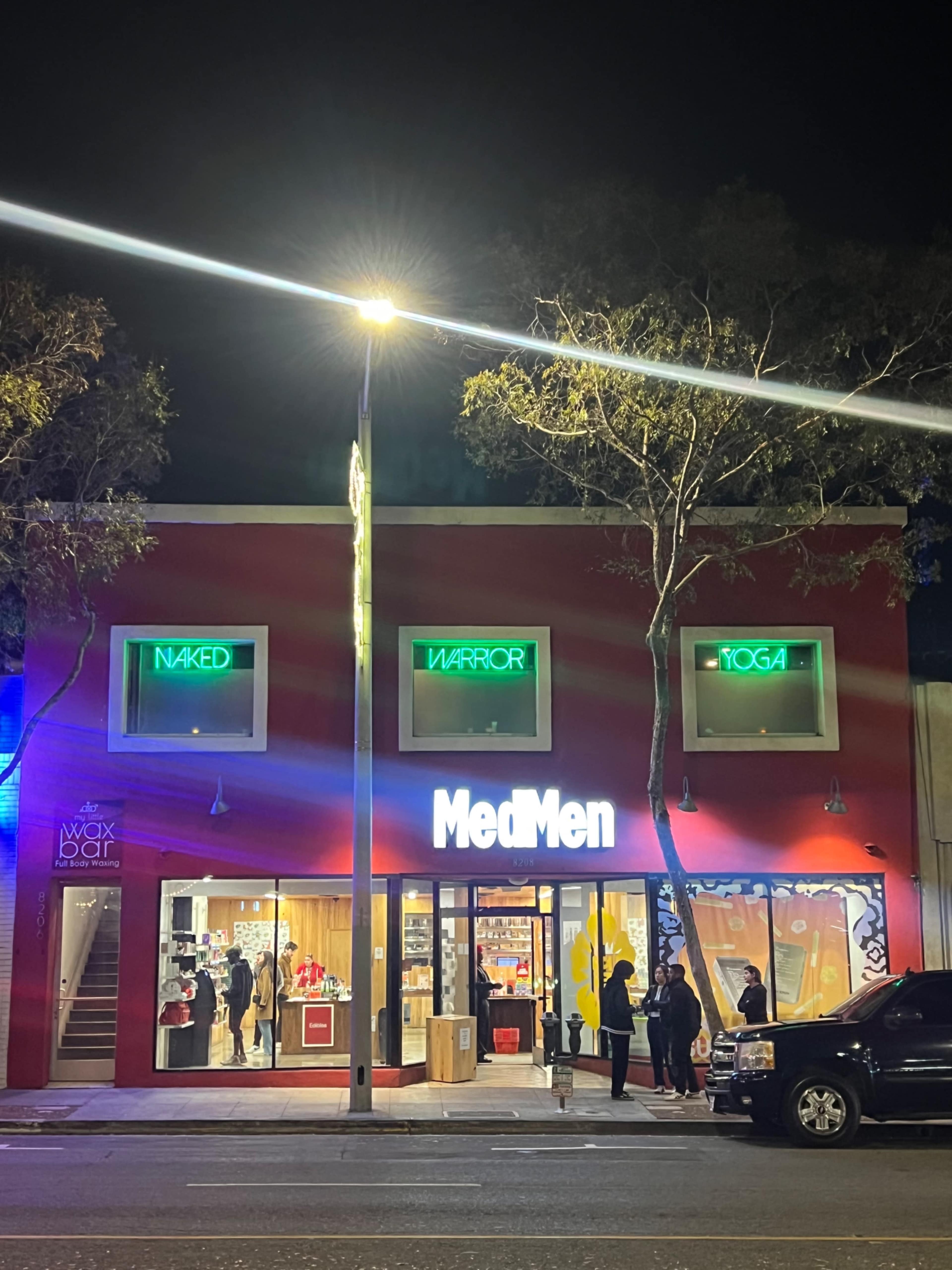The image shows a brightly lit storefront at night featuring the name "MedMen" prominently displayed, with other signs for "Wax Bar," "Naked," "Warrior," and "Yoga" visible in the background.