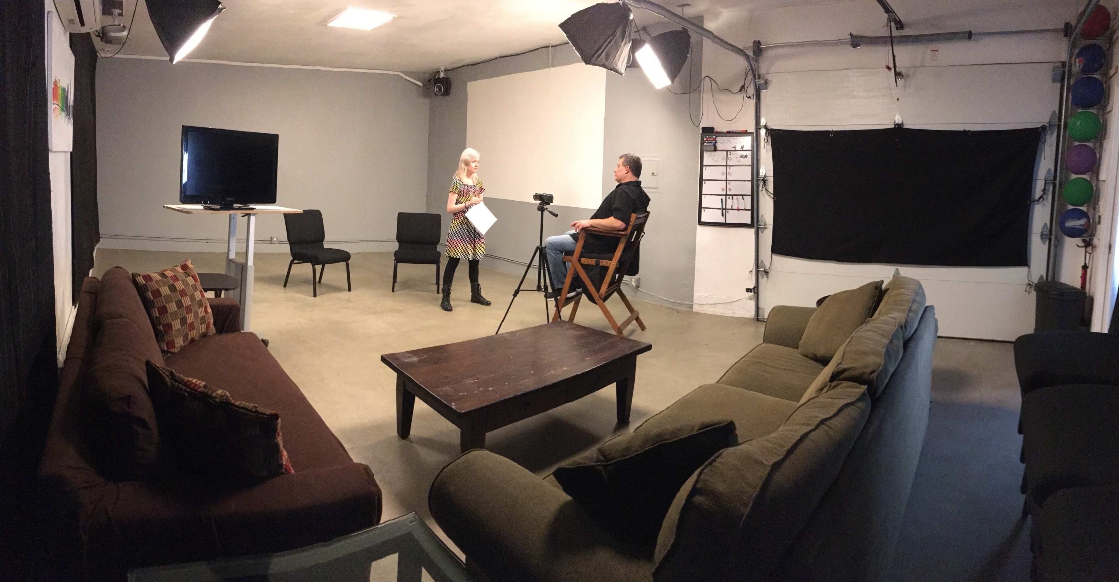 A woman stands holding a script while a man sits on a director's chair, both positioned in a studio set with chairs, a table, and soft lighting.