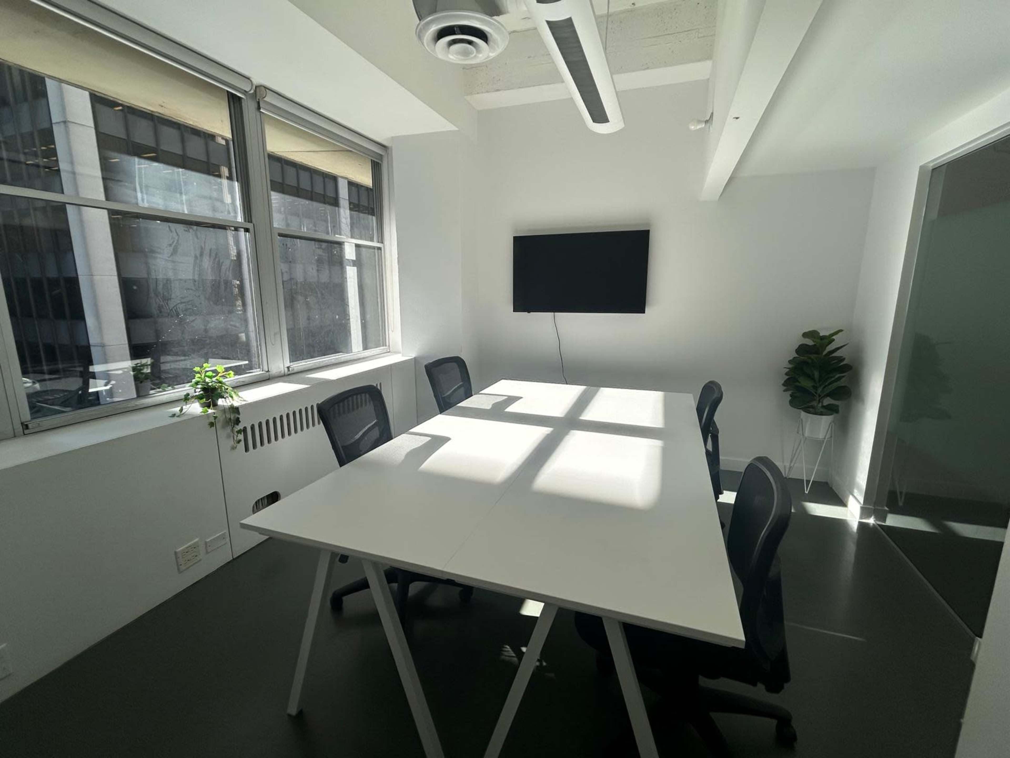 The image shows a bright, minimalistic conference room with a white table, black chairs, a wall-mounted television, and large windows allowing natural light to enter.