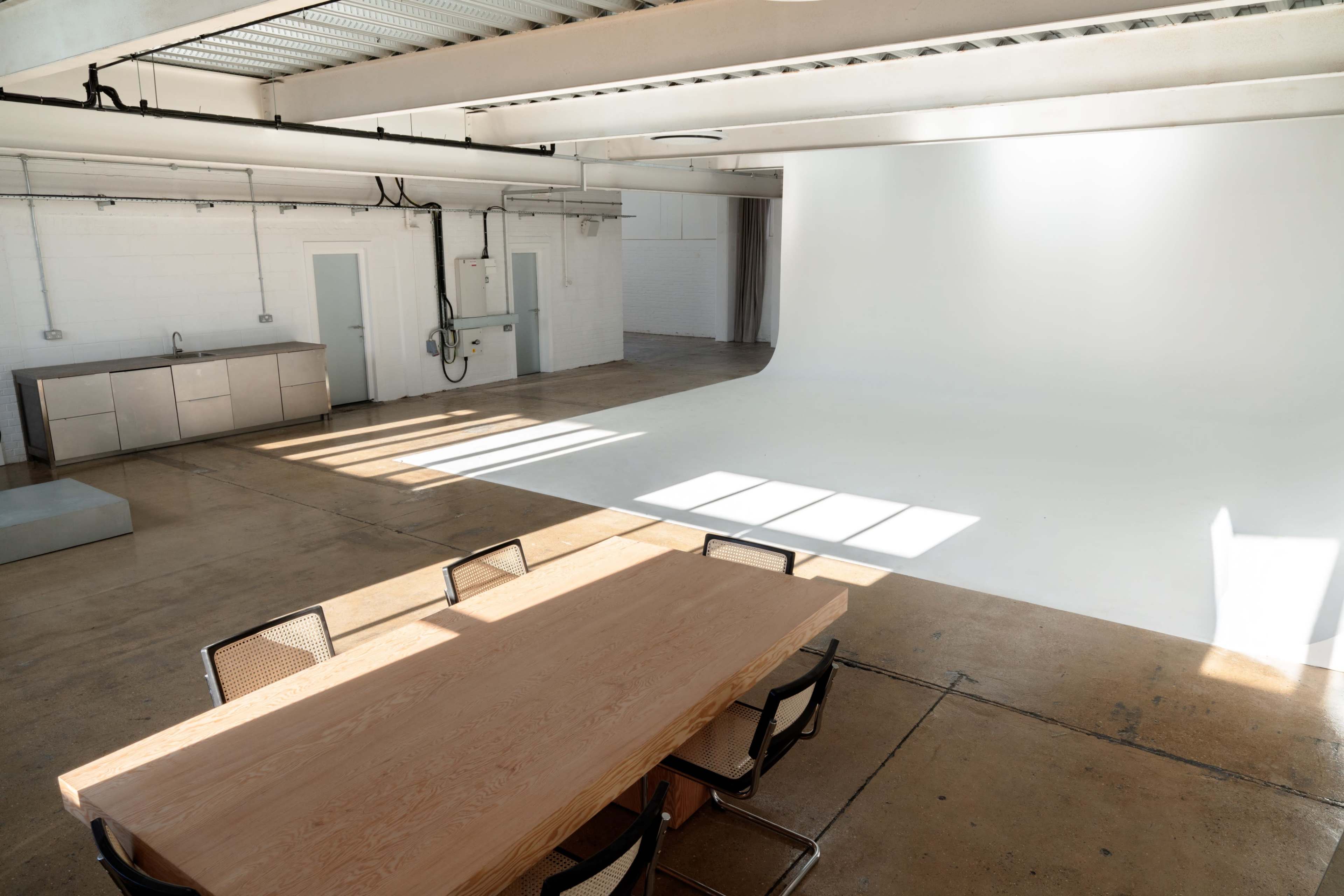 A spacious, well-lit studio features a long wooden table with several chairs and a large seamless white backdrop in the background.