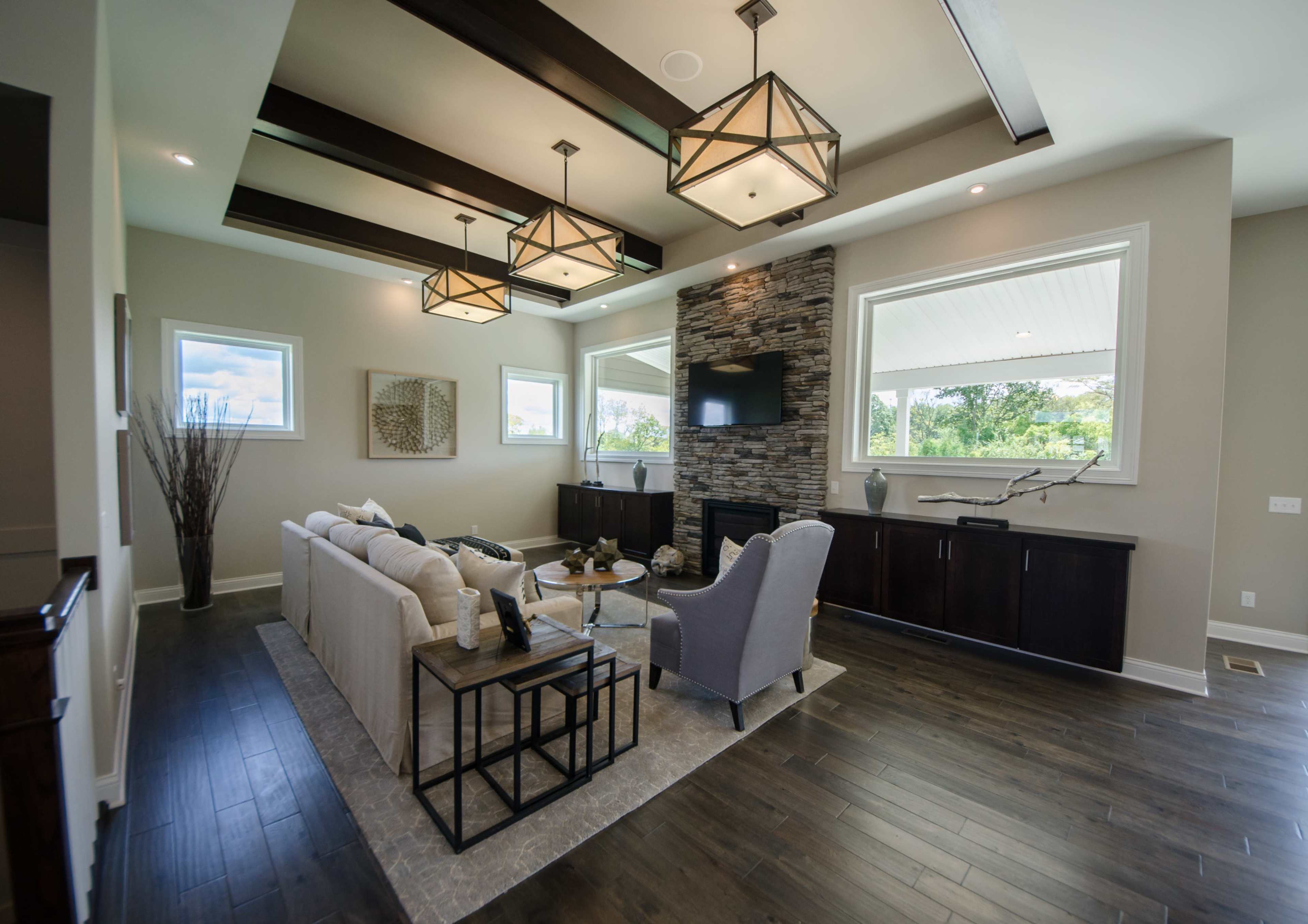 A modern living room features a stone fireplace, a sectional sofa, and wooden beams above, with large windows letting in natural light.