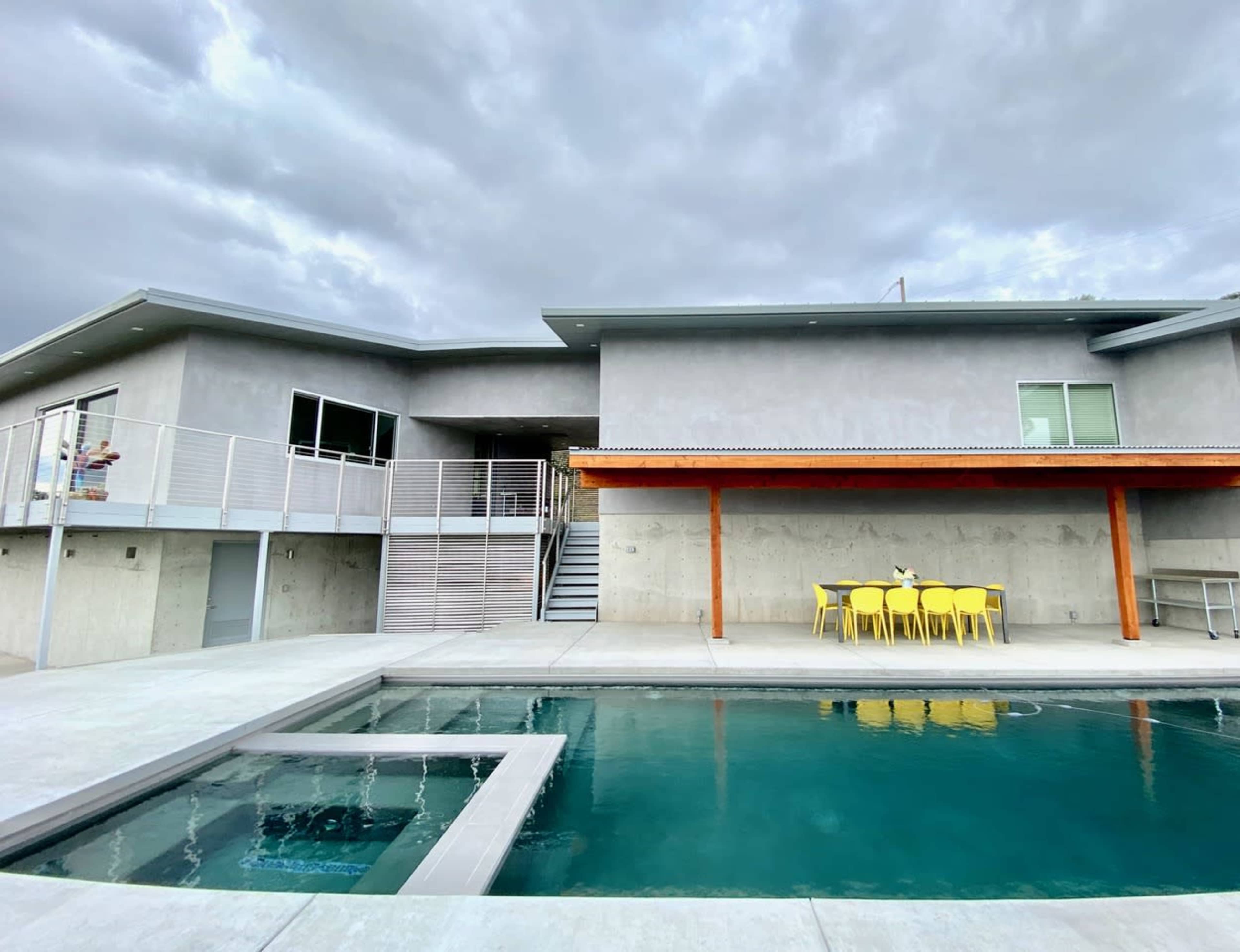 A modern house features a swimming pool and a patio with yellow chairs under an overhang, set against a cloudy sky.