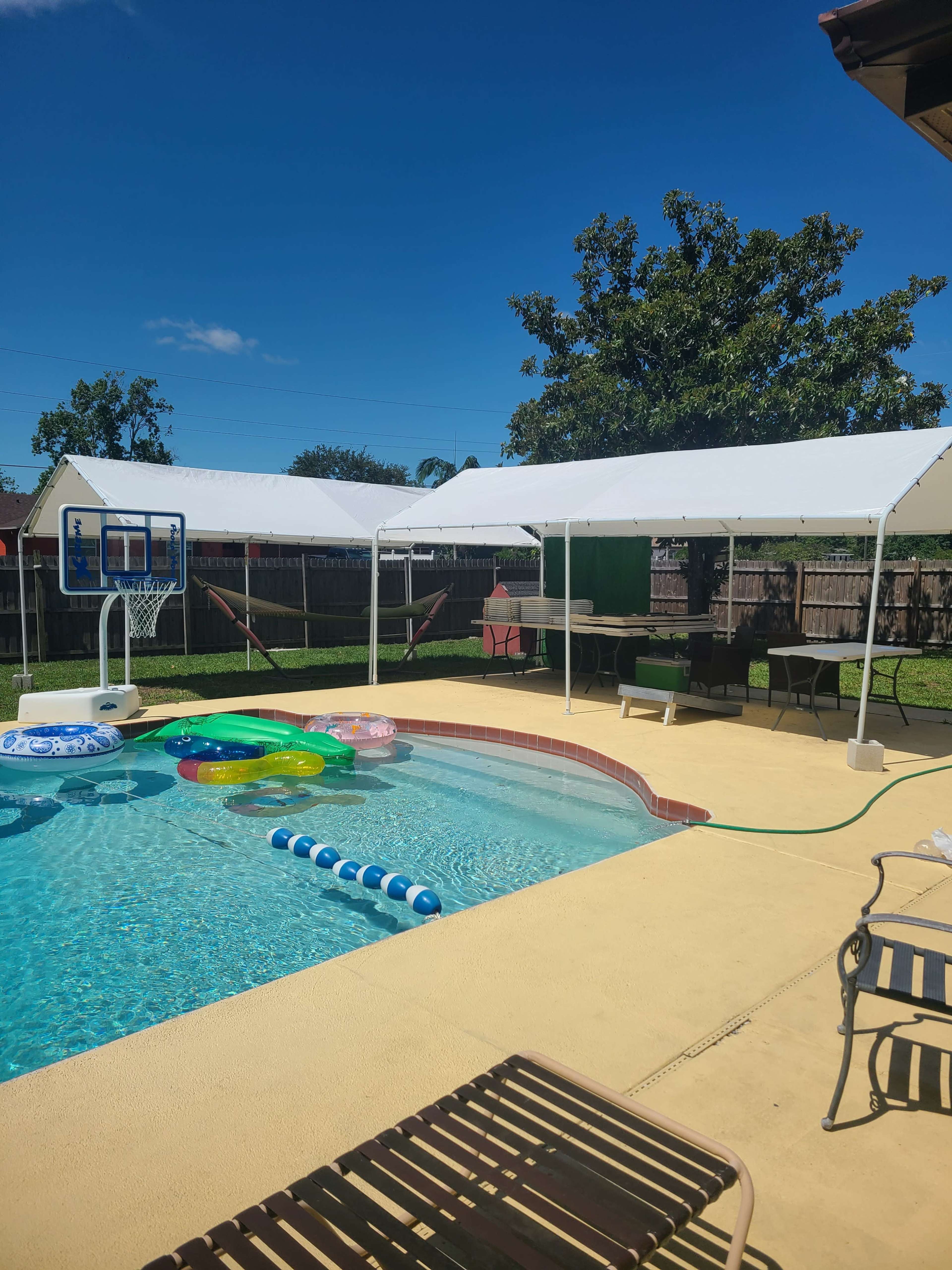 A backyard pool area with a basketball hoop, several floating toys in the water, and two large tents set up beside the pool.