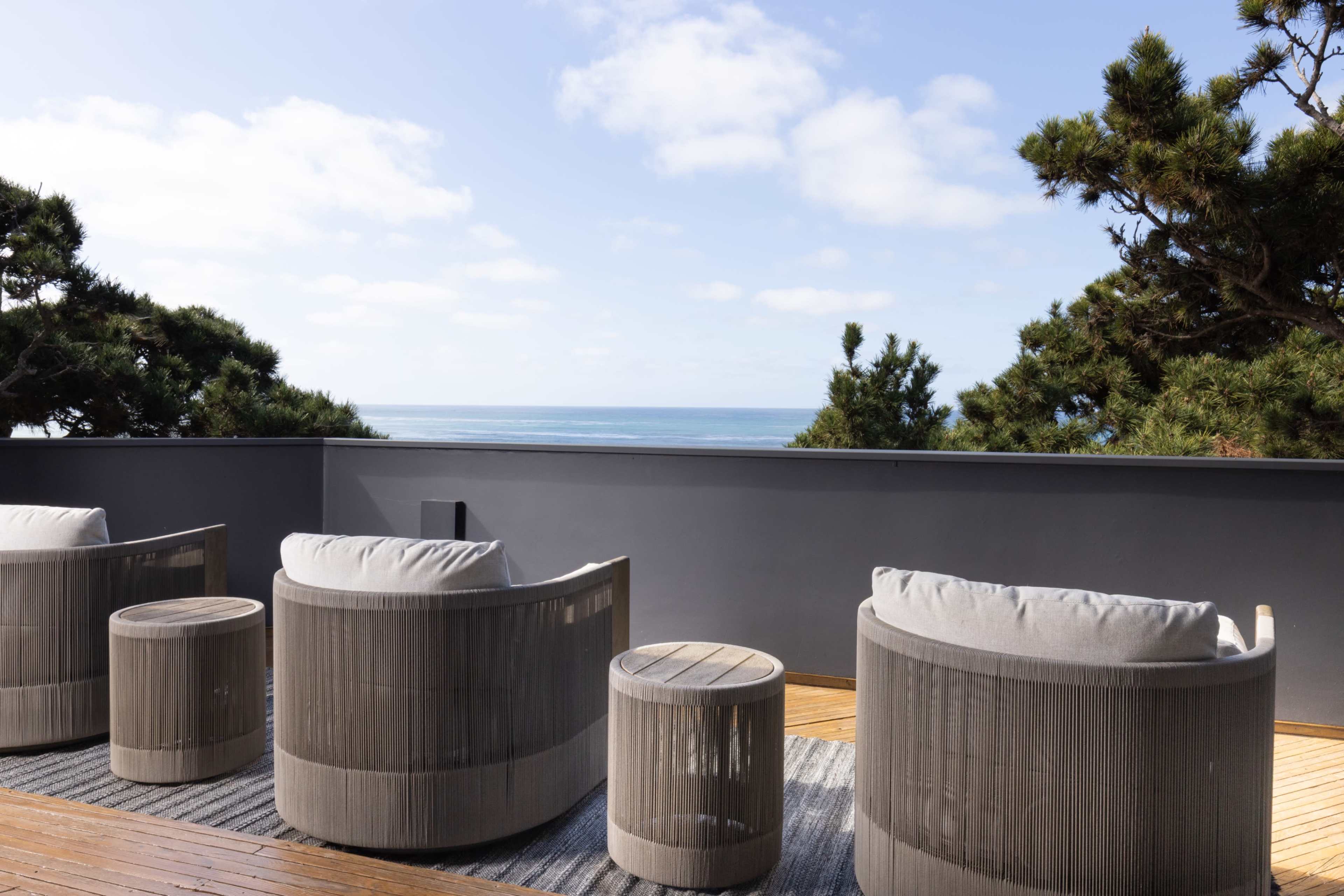 A coastal terrace features four circular chairs and a small table, overlooking the ocean under a clear blue sky.