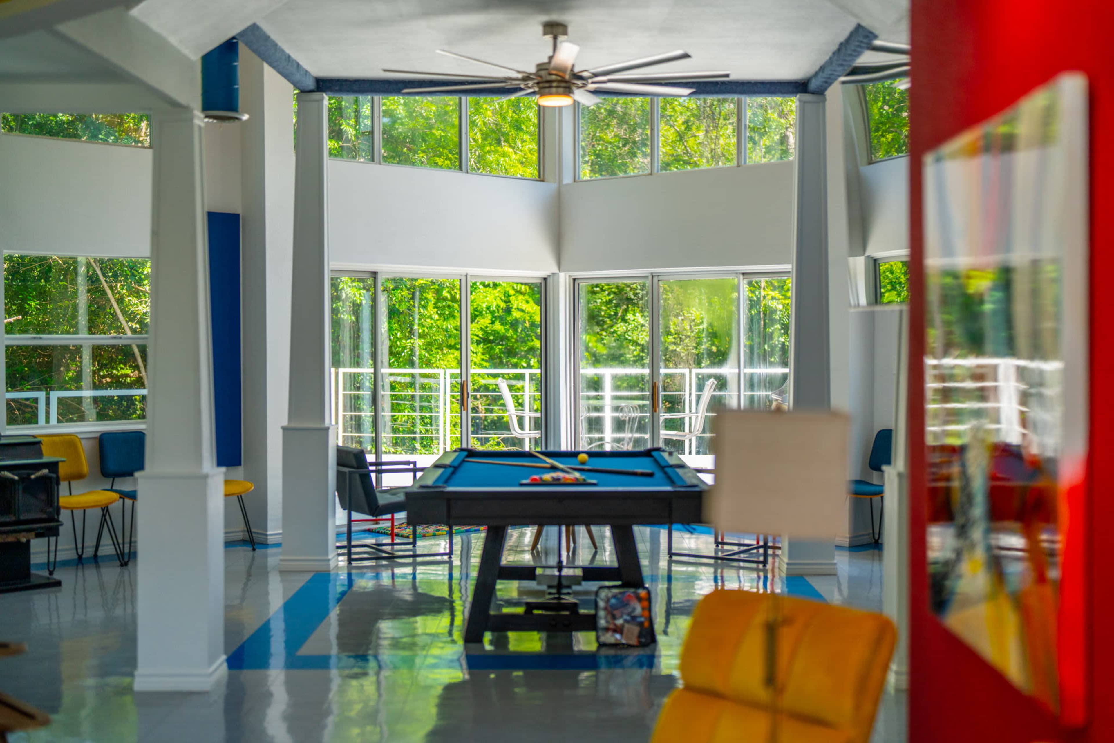 A brightly lit game room with large windows, featuring a pool table at its center and modern seating around the space.