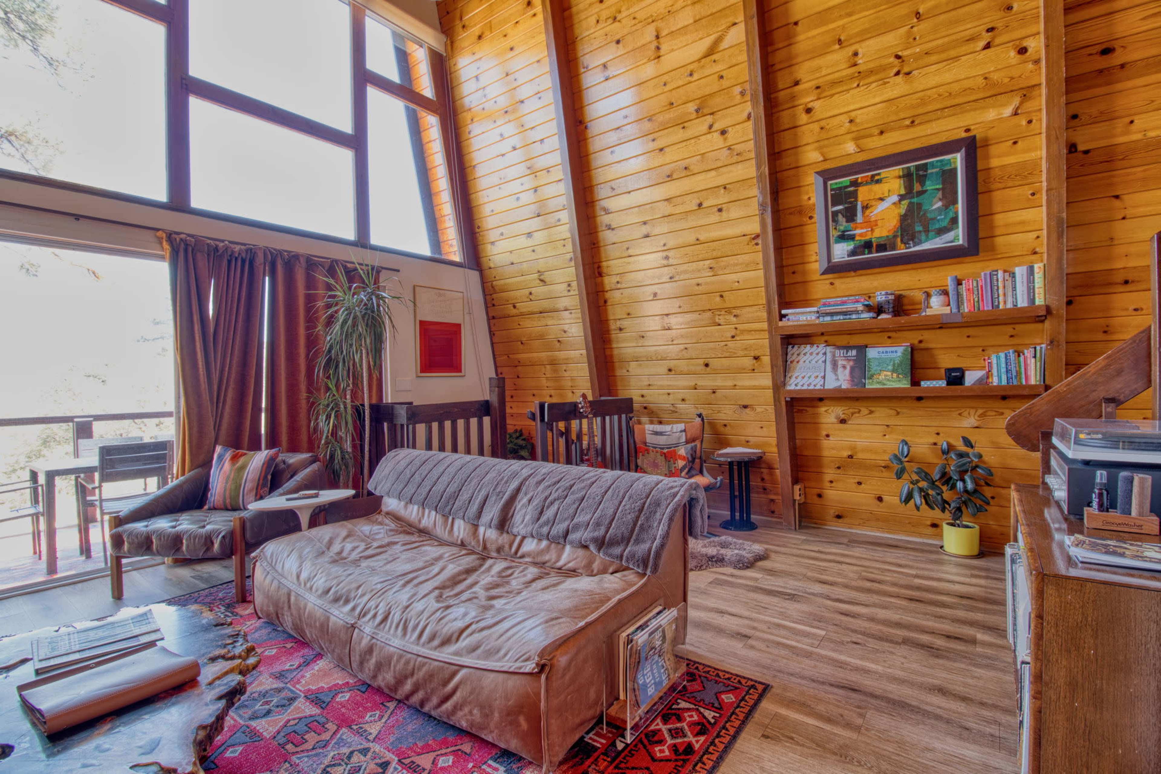 The image shows a cozy living room with wooden walls, a large window letting in light, and a mix of furniture including a sofa, chair, and bookshelves.