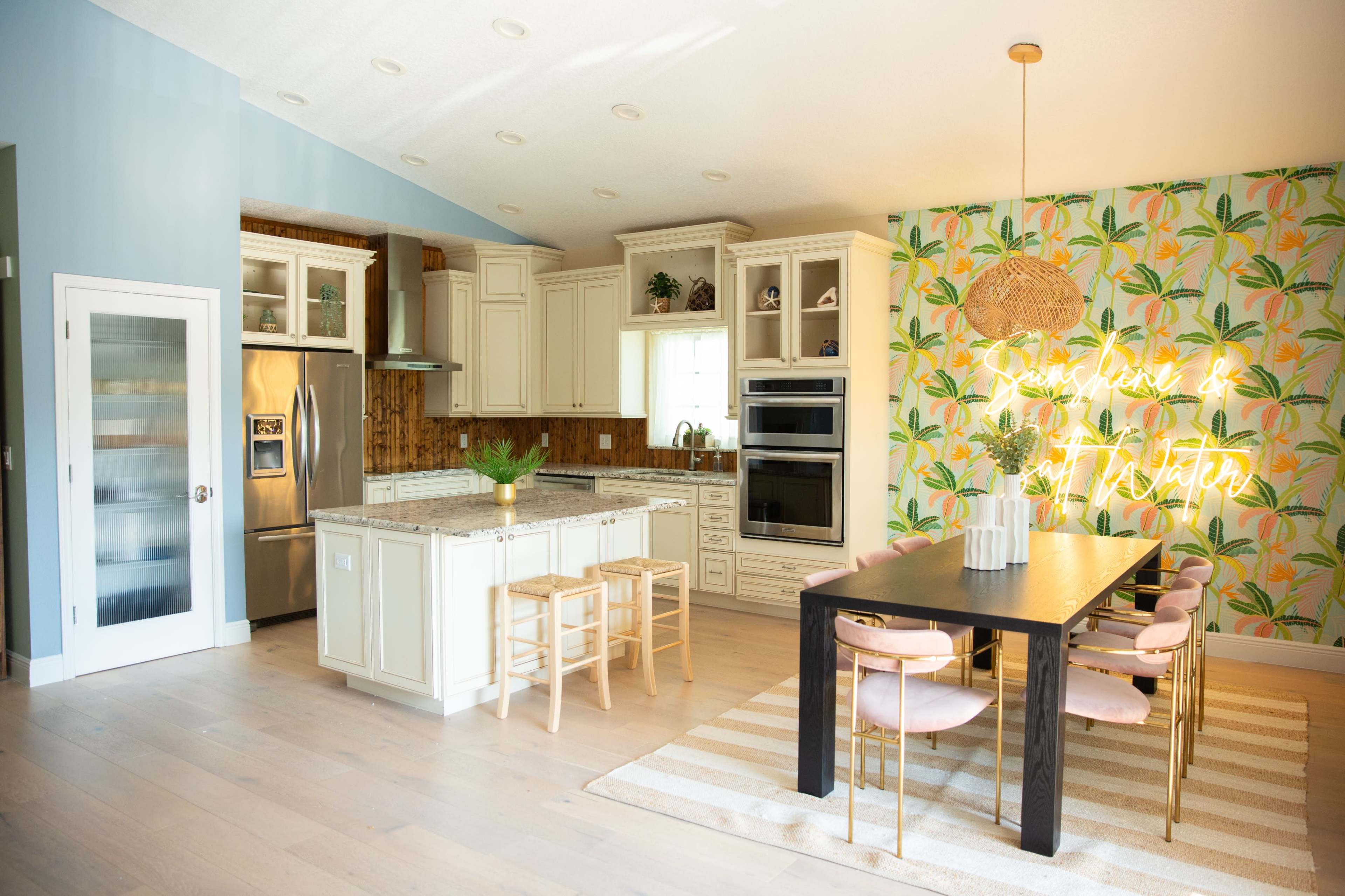 The image shows a modern kitchen with light cabinets, stainless steel appliances, and a dining area featuring a black table and pink chairs, set against a tropical wallpaper and neon sign.