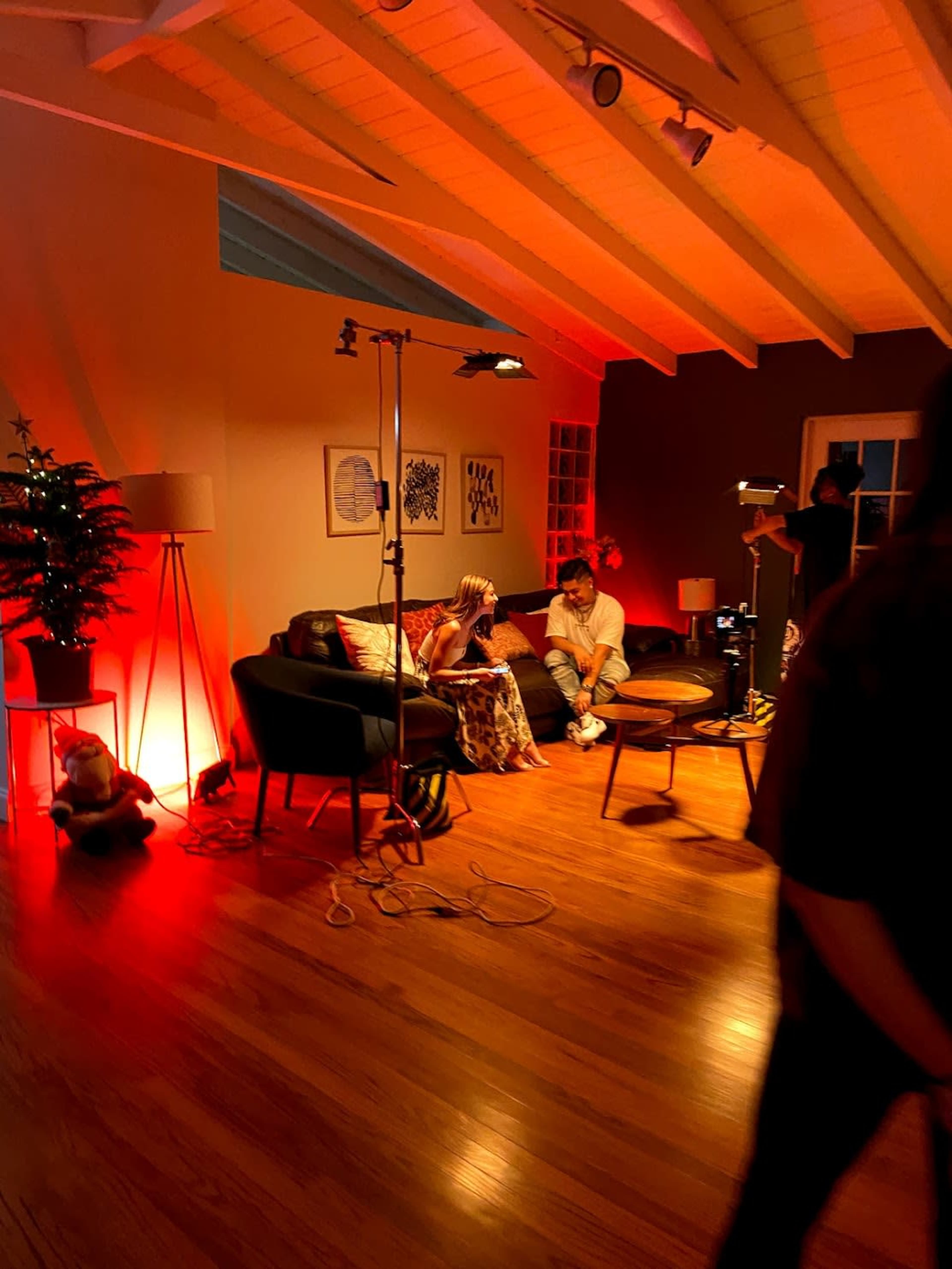 The scene depicts a cozy room with two individuals seated on a couch under warm red lighting, surrounded by various lighting equipment and a decorative tree.