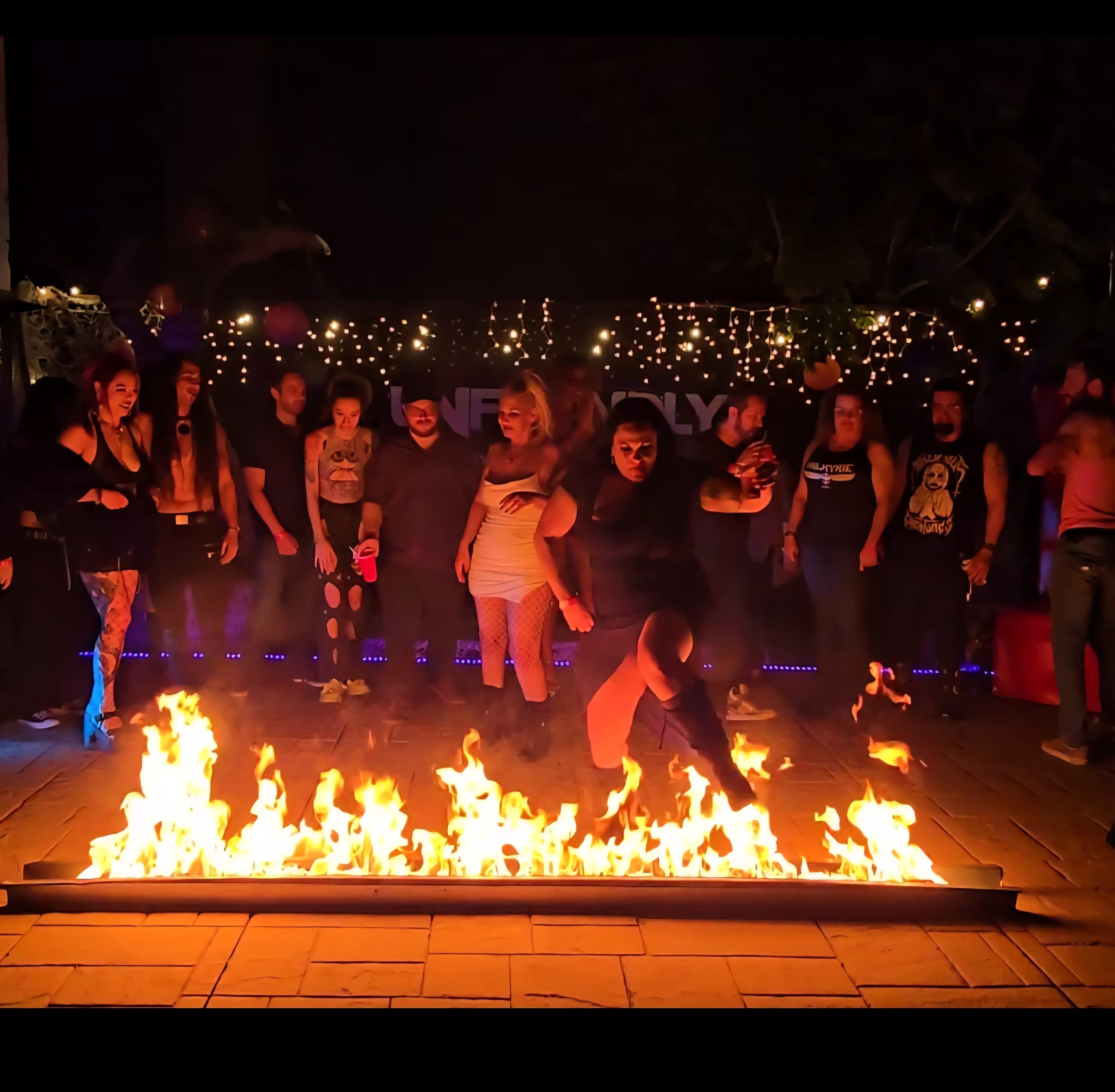 A group of people stands around a fire performance at night, watching as one performer dances near the flames in front of a festively decorated backdrop.