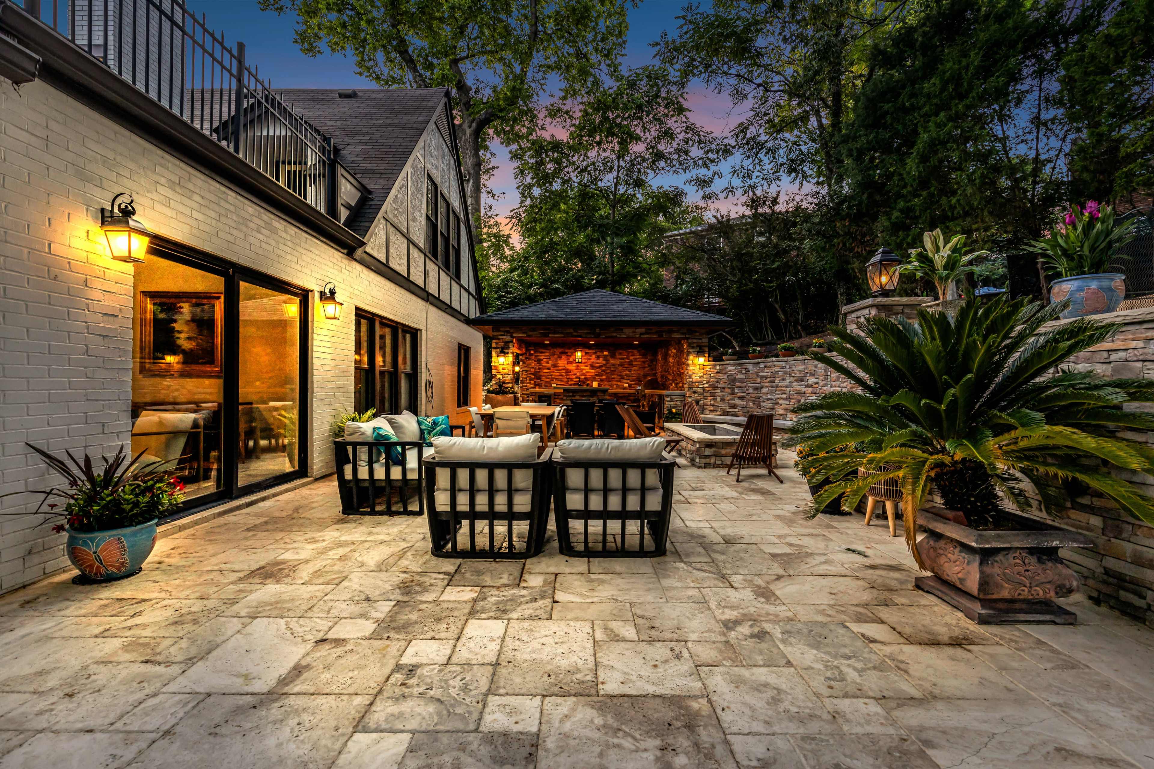 The image shows a stone patio area with seating arranged around a fire pit and a gazebo, set against a backdrop of a two-story house surrounded by greenery at dusk.