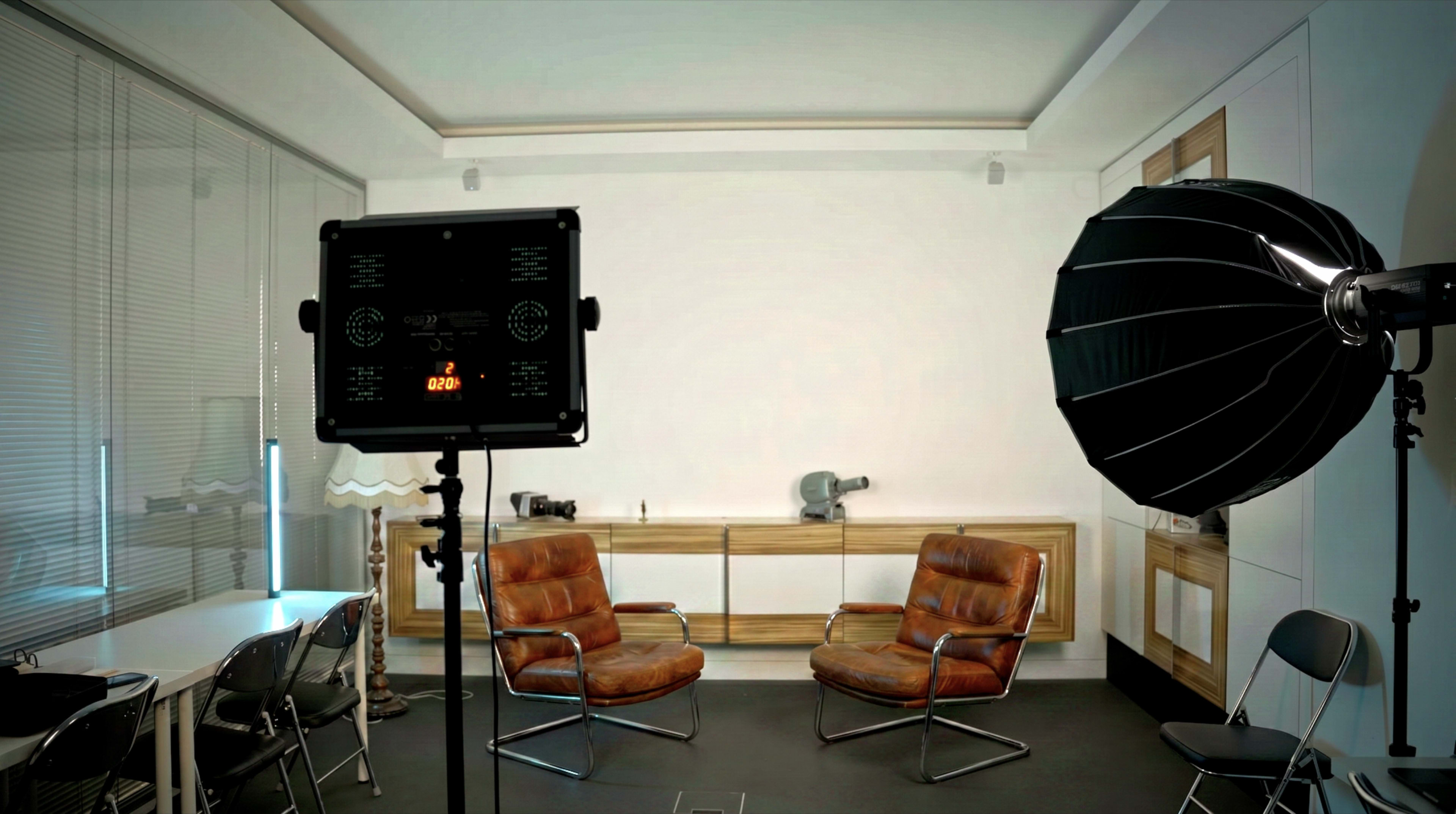 The image shows a minimalist room with two brown leather chairs, a lighting setup, and a backdrop of wooden cabinetry.
