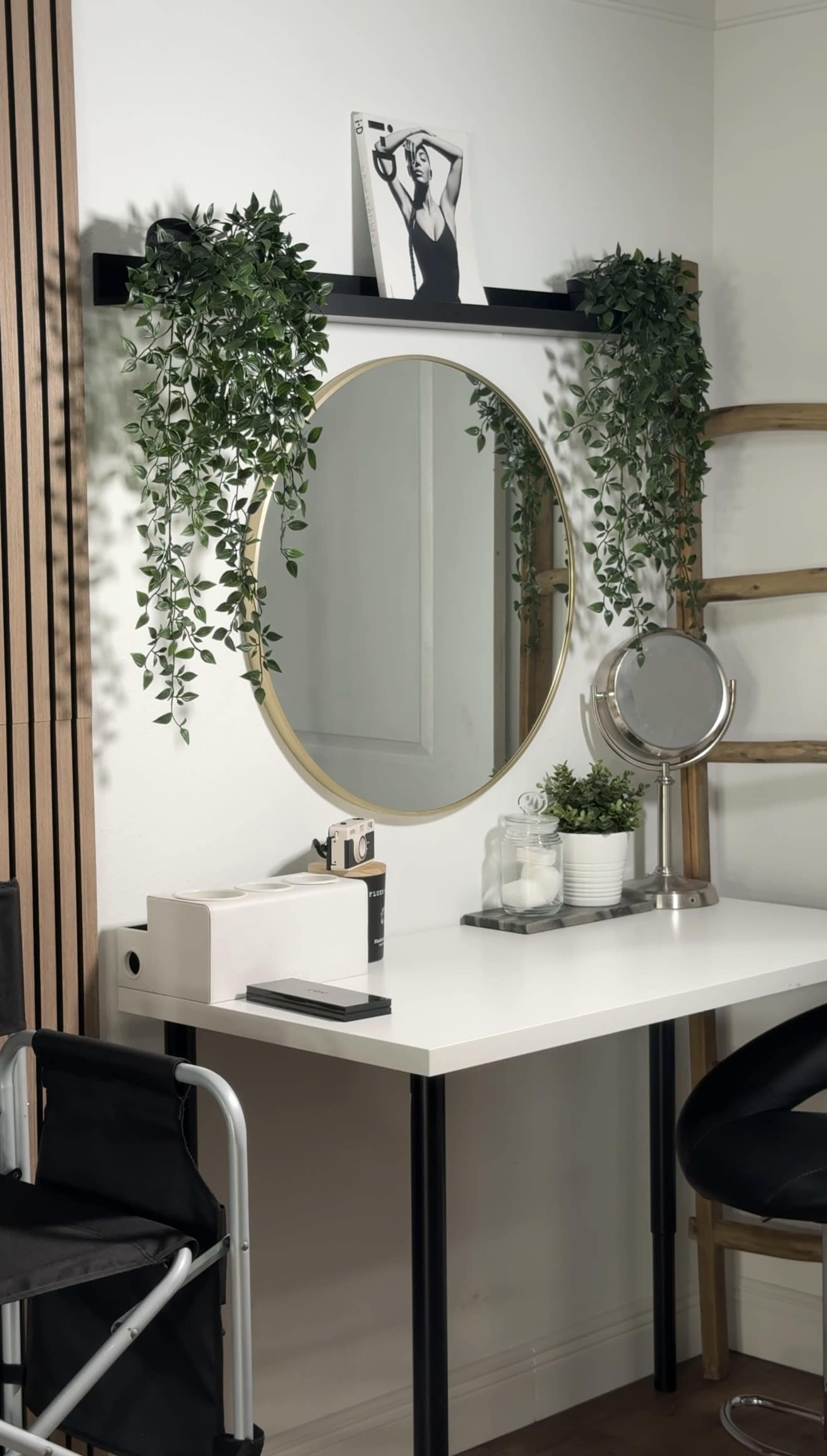 A modern vanity table features a large round mirror, potted plants, and a small shelf with a magazine against a minimalist background.