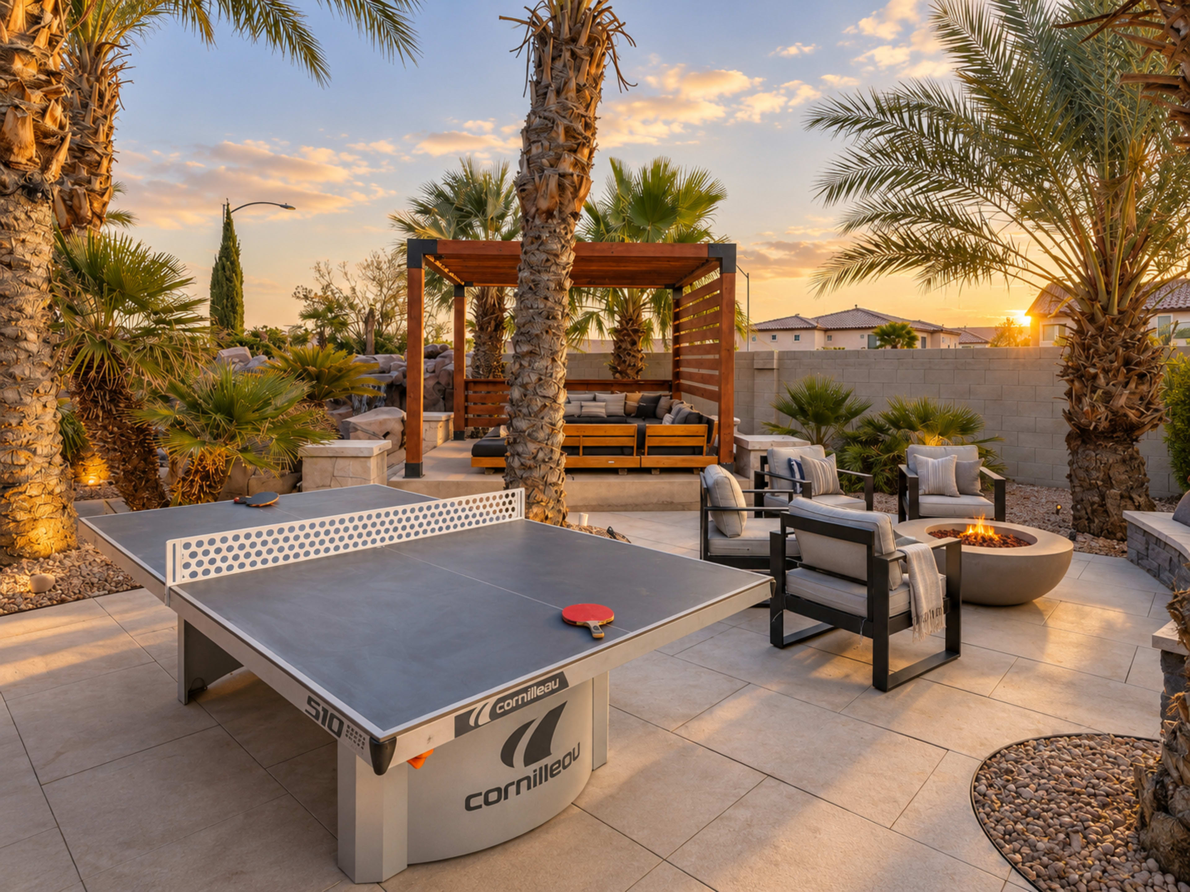 The image shows a backyard patio featuring a ping pong table, surrounded by palm trees, modern outdoor furniture, and a fire pit, with a sunset casting warm light across the scene.
