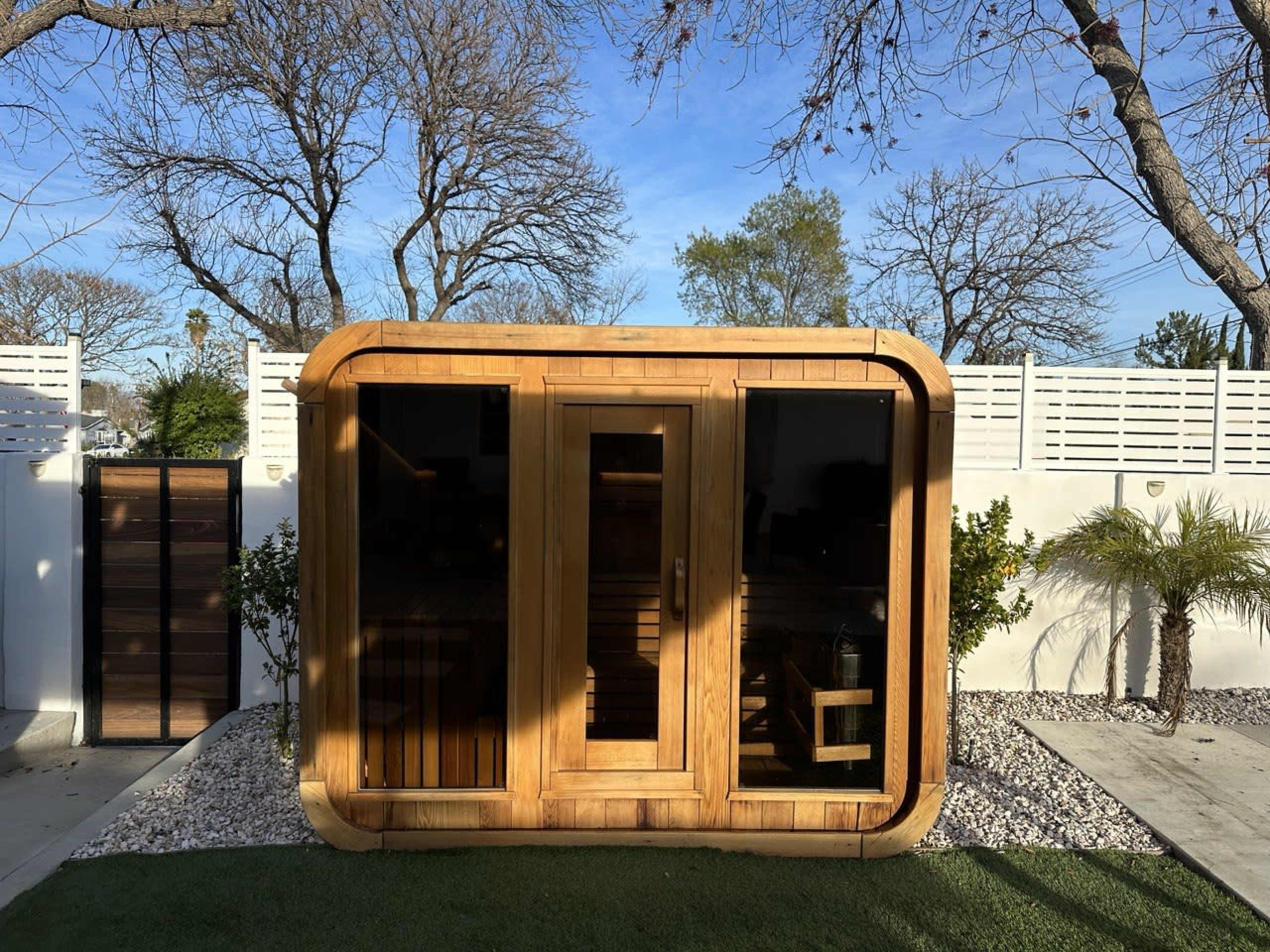 A modern wooden sauna structure with large glass panels, surrounded by trees and landscaped gravel.