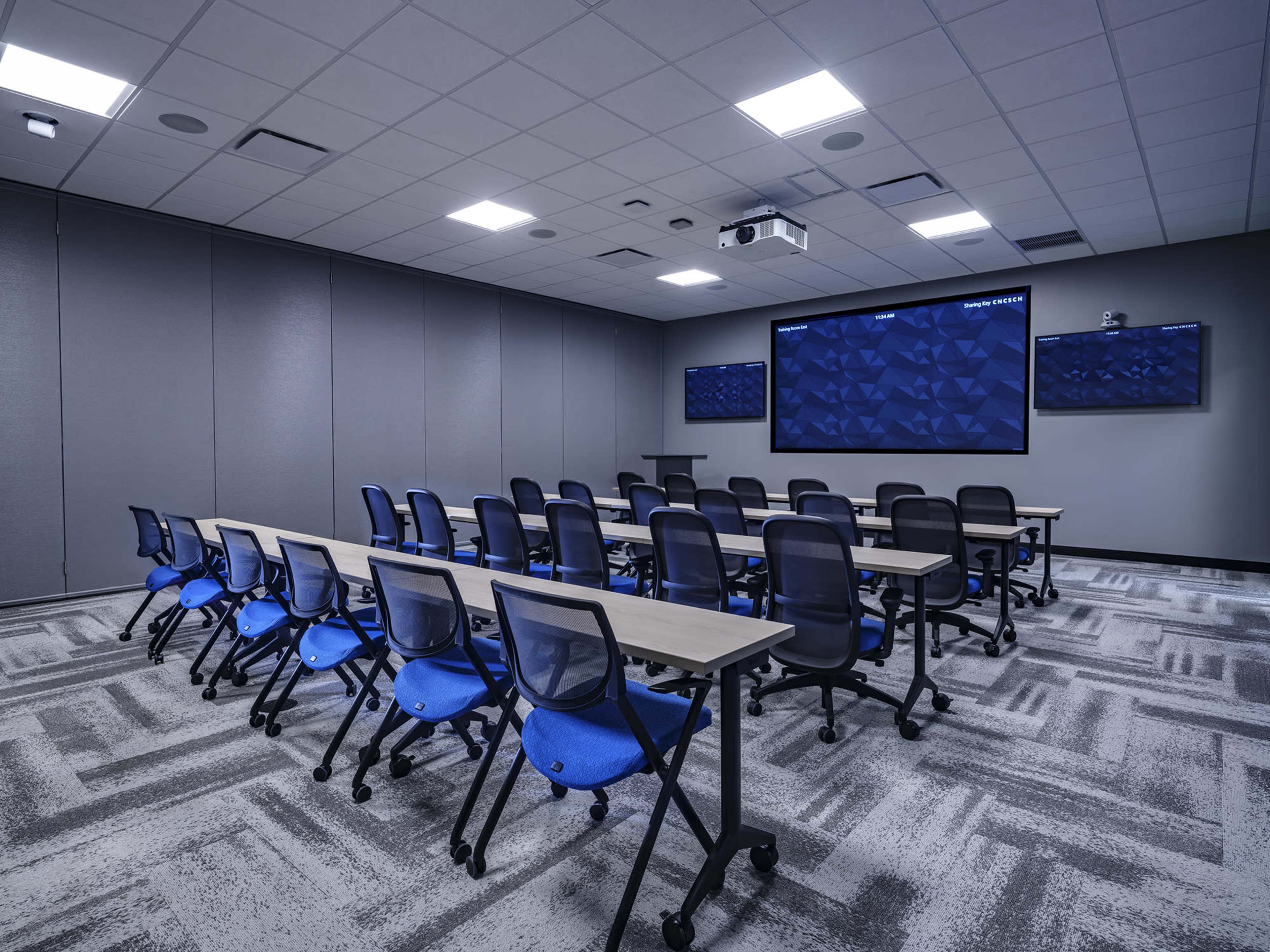 The image shows a modern conference room with several rows of chairs and tables, featuring two large screens on the front wall.