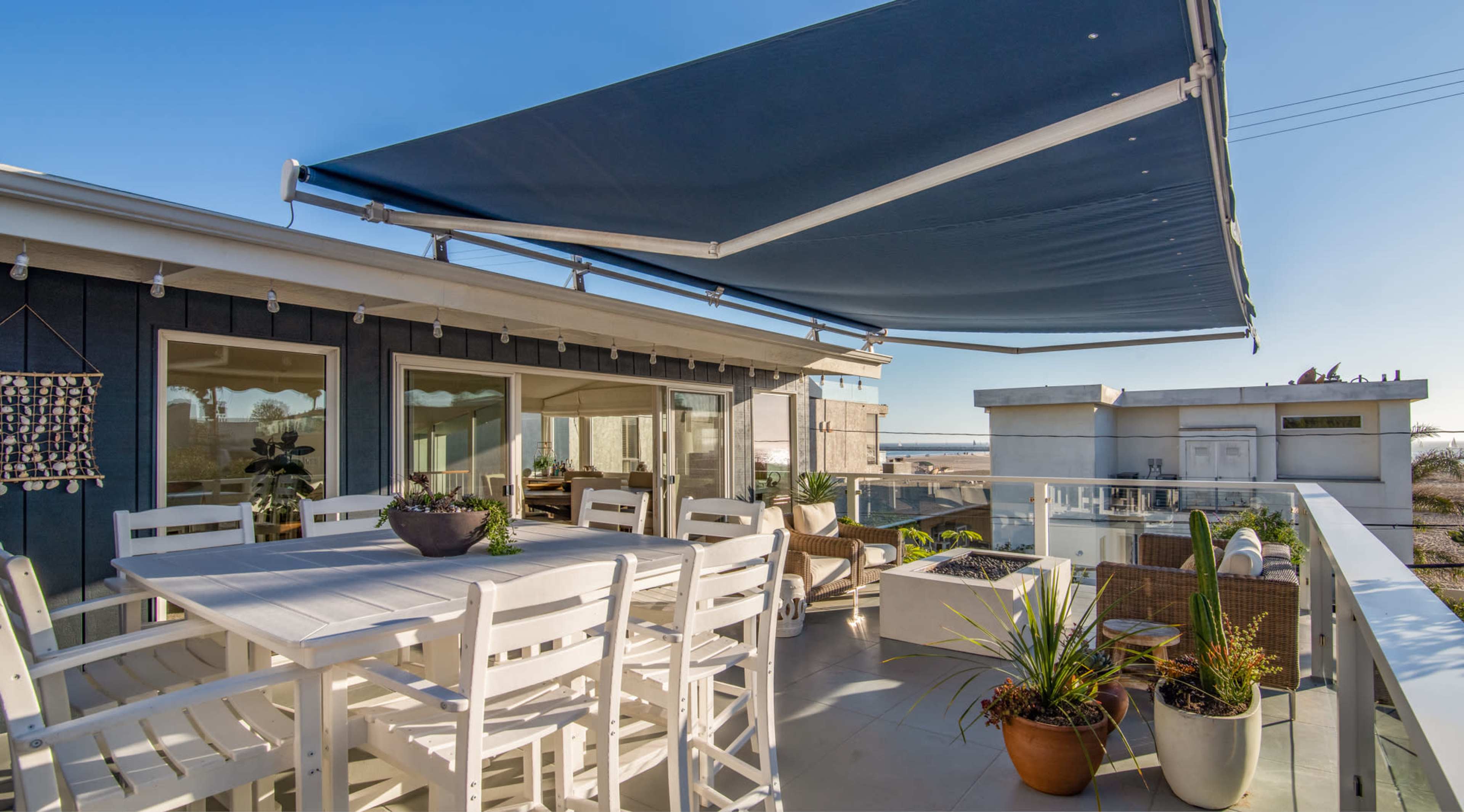 A rooftop terrace featuring a large white dining table surrounded by chairs, overlooked by a blue retractable awning, with decorative plants and a view of a coastal landscape.