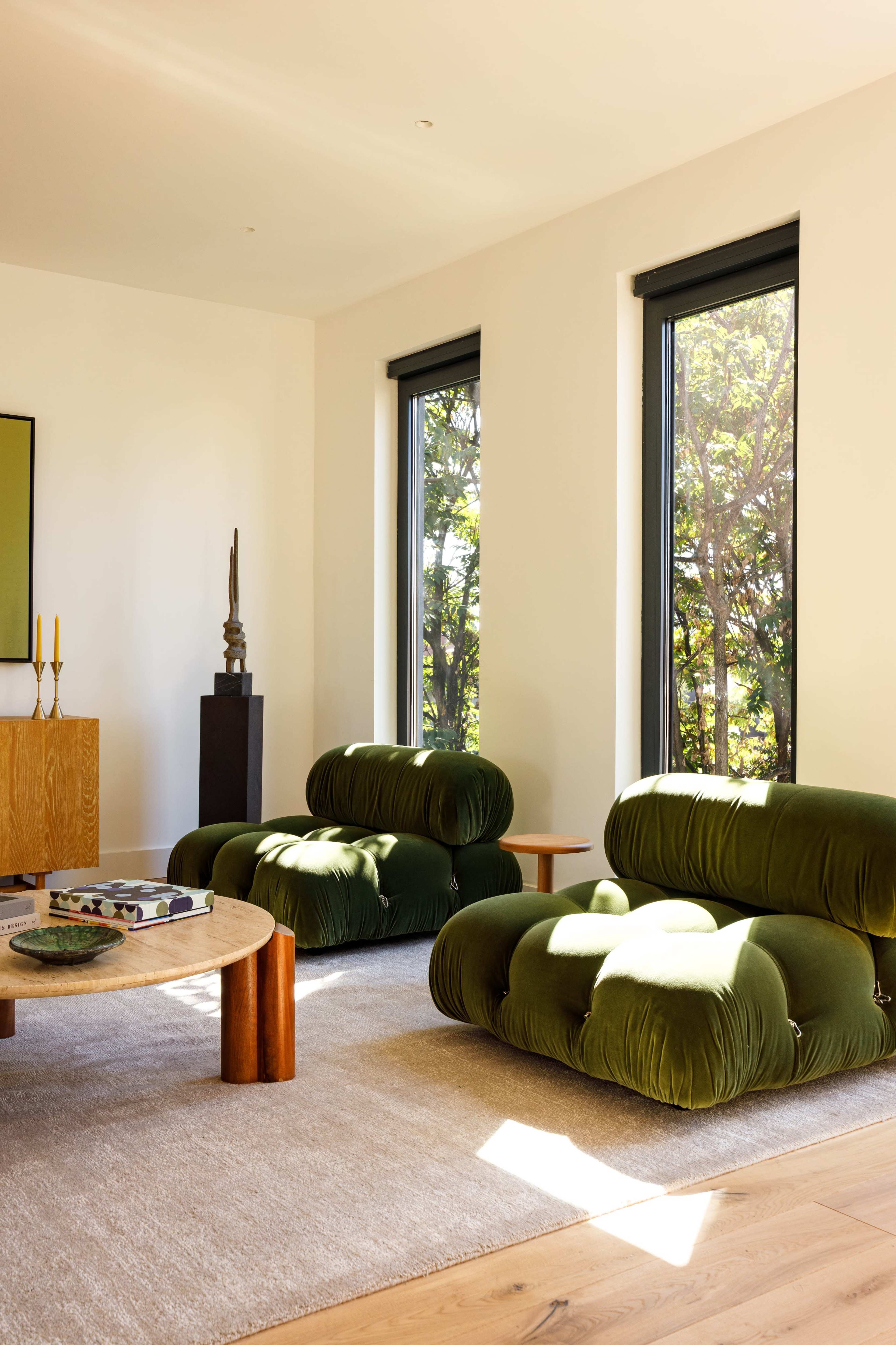 The image features a modern living room with two green, plush sofas, a round wooden coffee table, and large windows allowing natural light to enter the space.