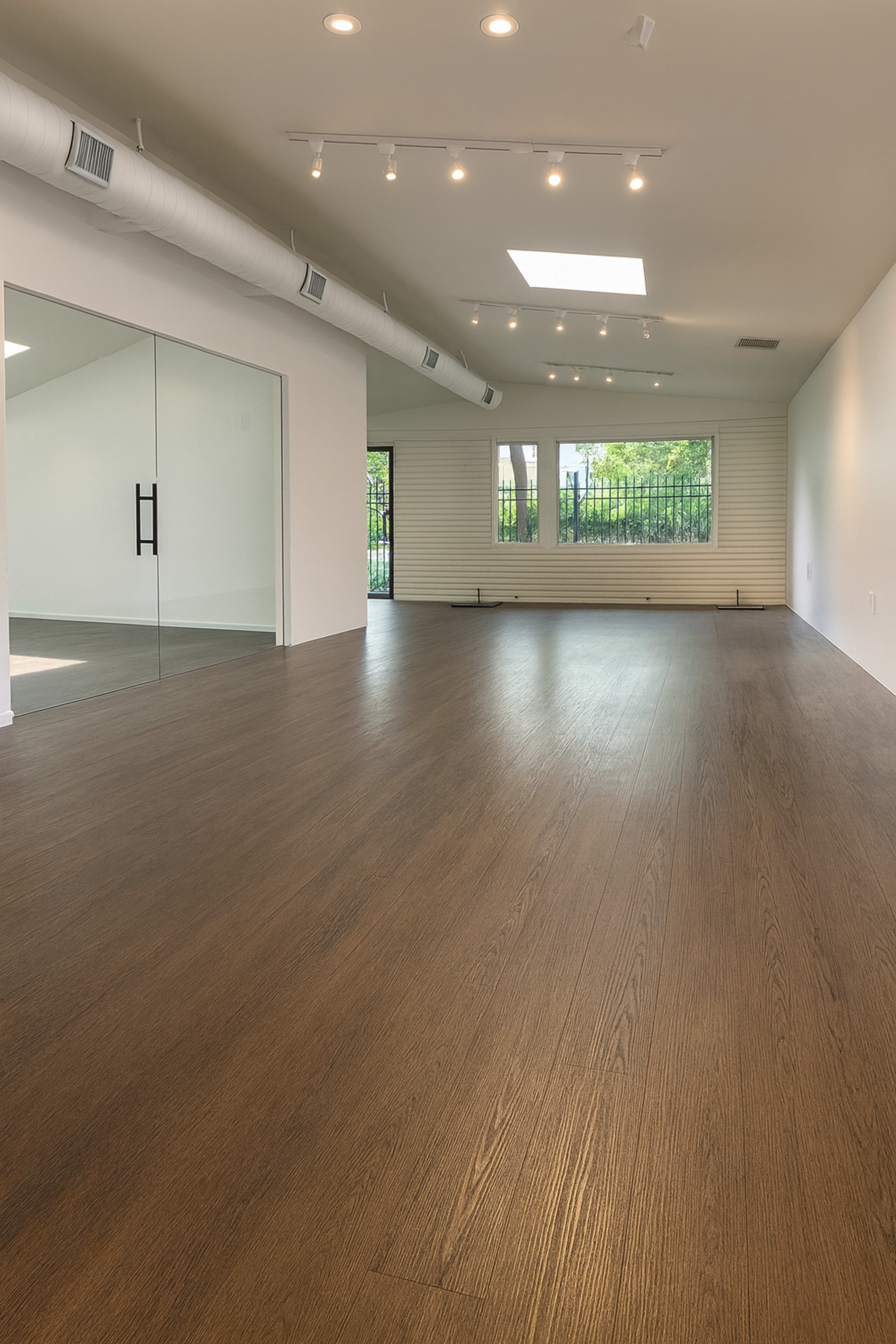 The image shows a spacious, empty room with hardwood flooring, large windows, and a skylight.