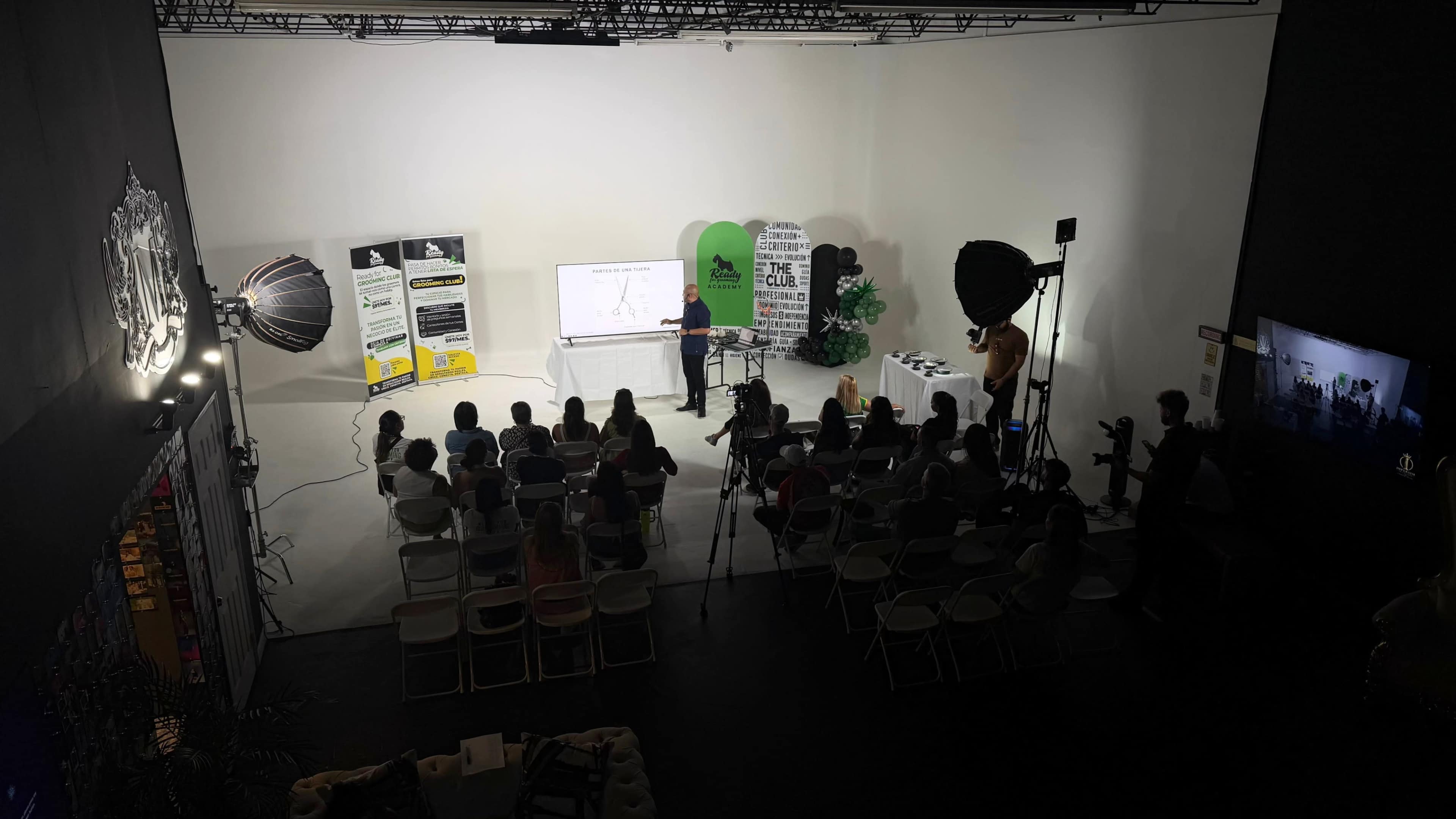 A presenter stands in front of a screen while an audience seated in white chairs observes in a well-lit studio.