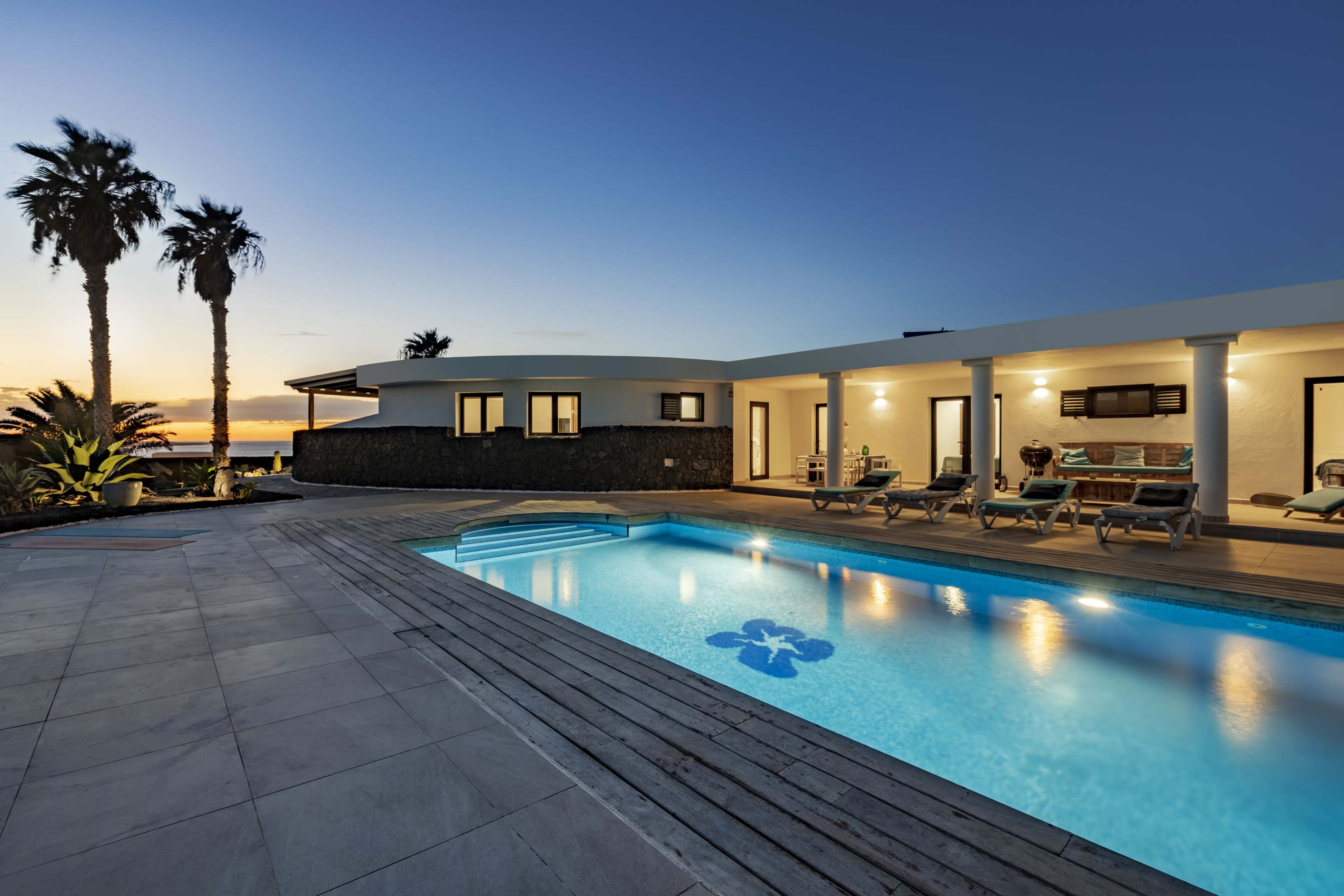A modern villa with a swimming pool and palm trees is illuminated by a sunset sky.