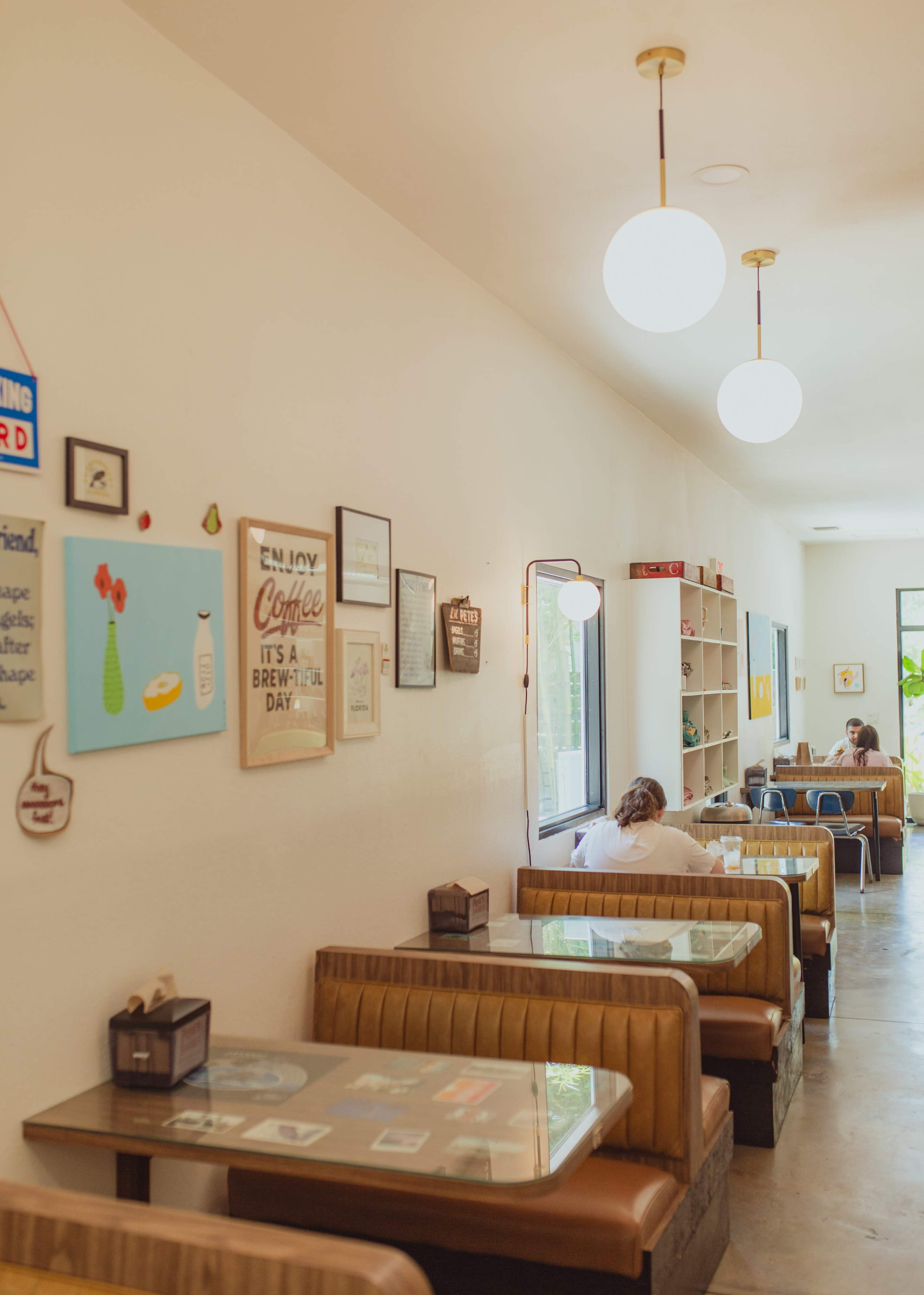 The image shows a bright, minimalist café interior with wooden booths, artwork on the walls, and a few patrons seated at tables.