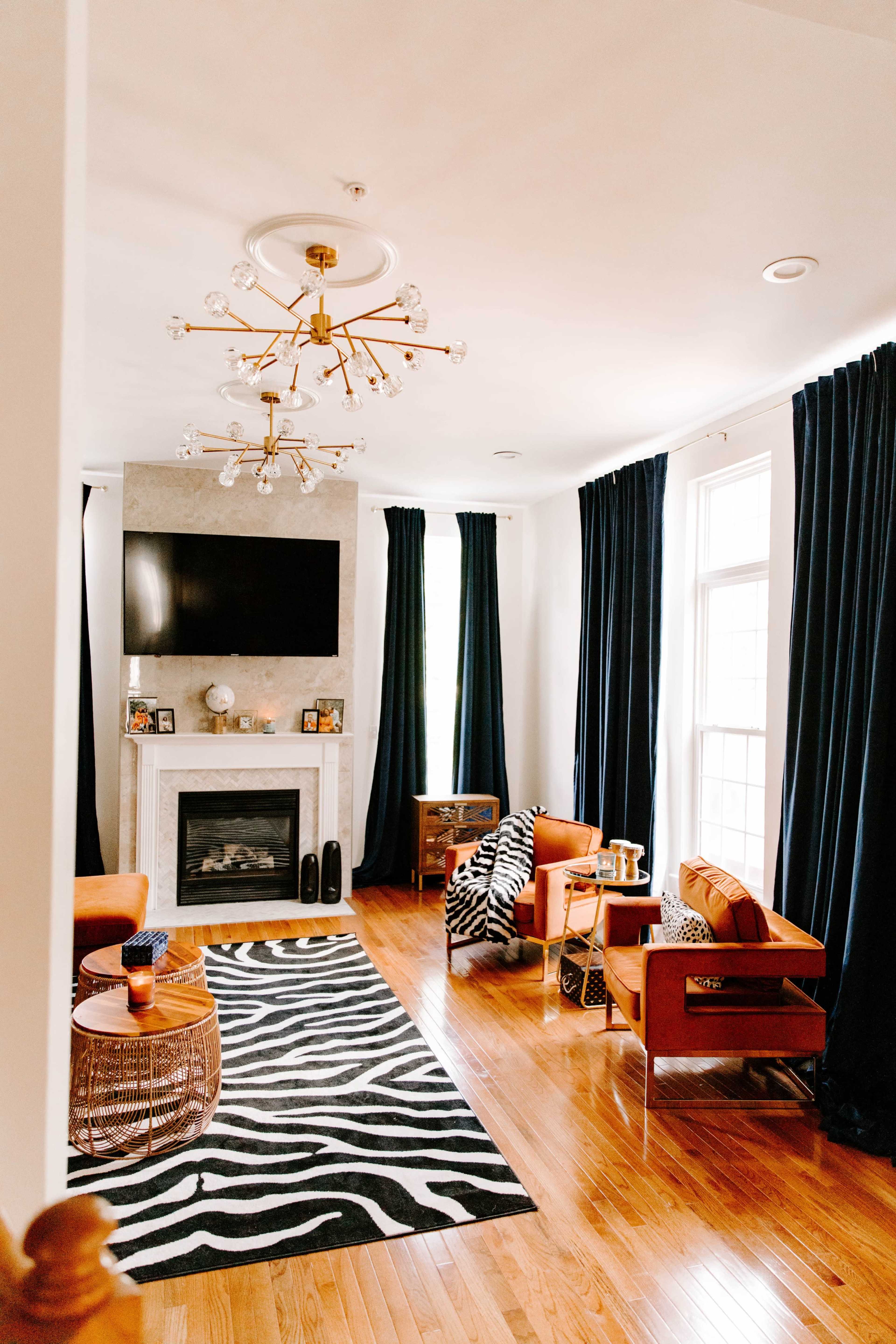 The living room features a fireplace with a TV above it, orange chairs, a zebra-patterned rug, and floor-to-ceiling dark curtains.