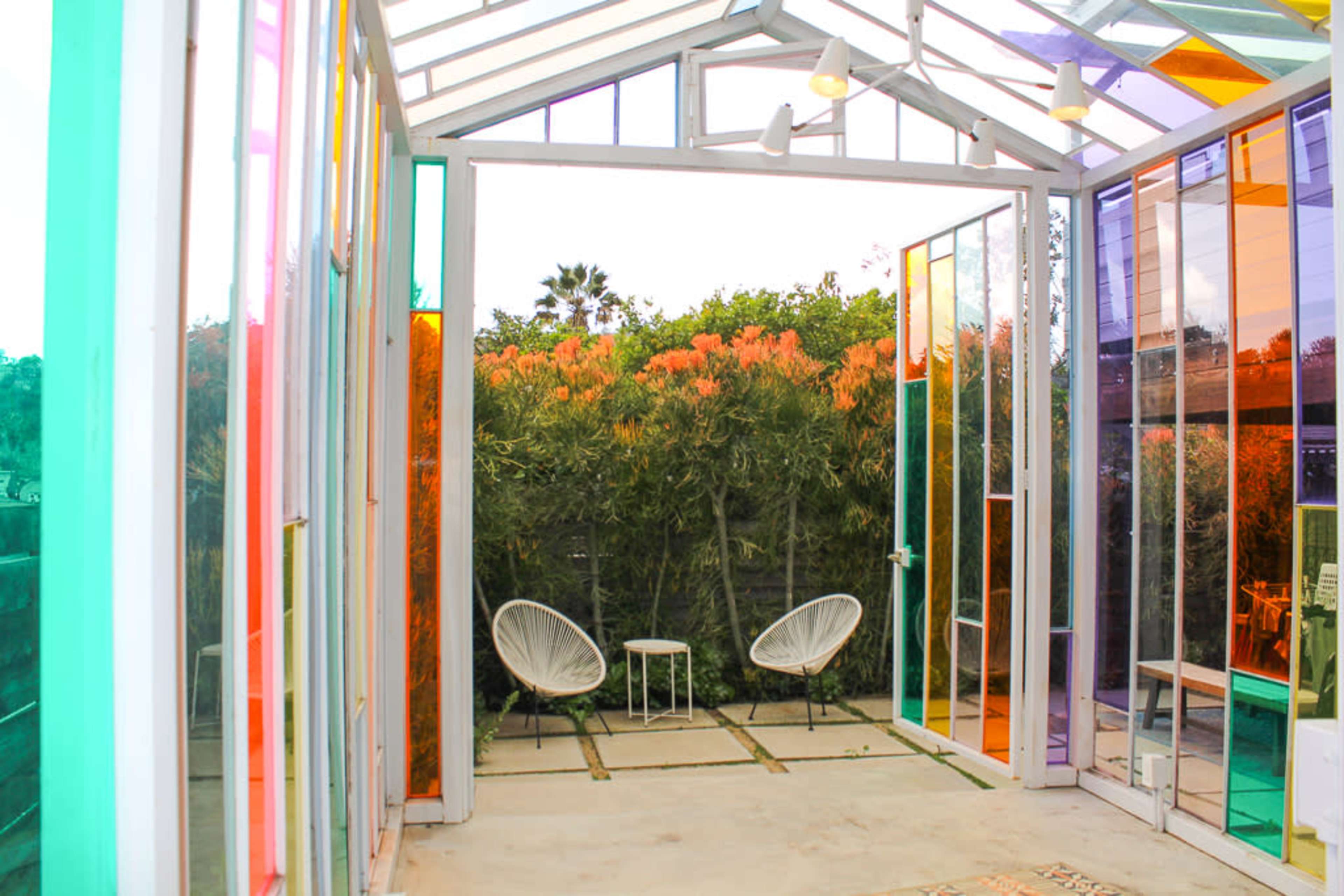The image shows a brightly colored glass room with two white chairs facing a garden filled with greenery and colorful plants.