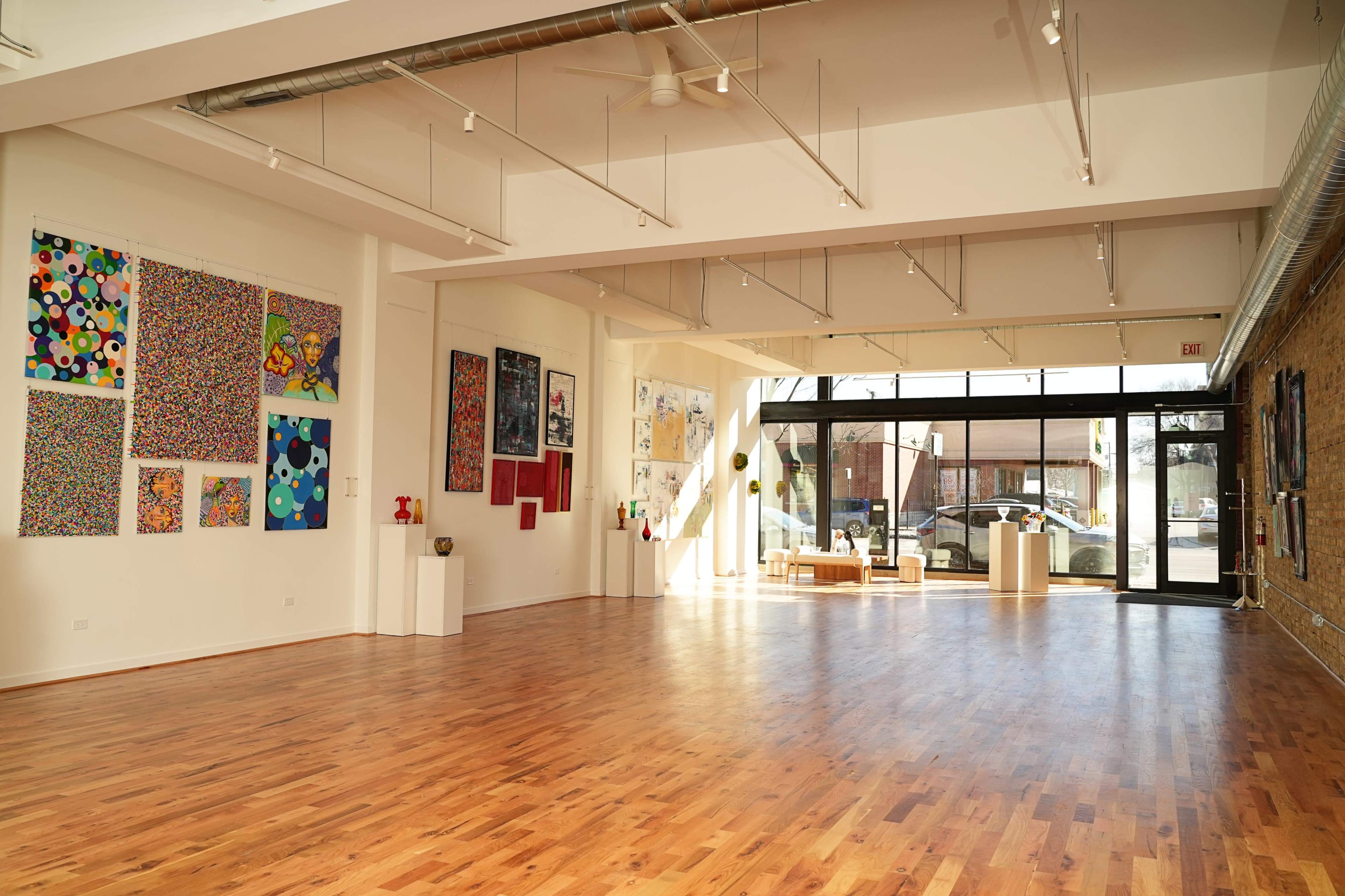 The image shows a spacious art gallery with a wooden floor, featuring colorful contemporary artwork on the walls and large windows allowing natural light to enter.