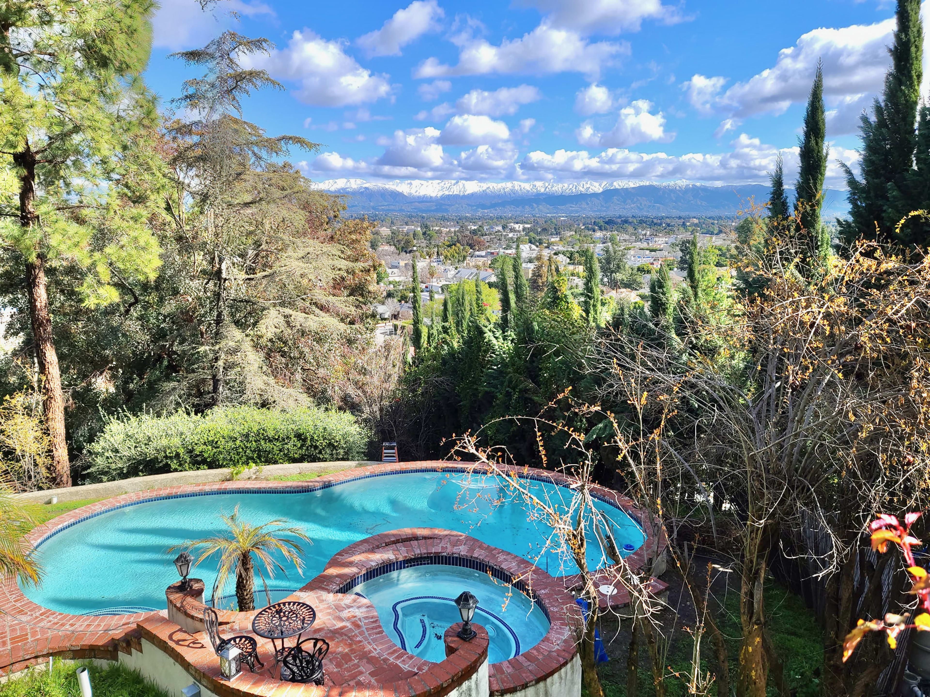 A swimming pool with a spa area, surrounded by trees and overlooking a mountainous landscape with a clear sky.