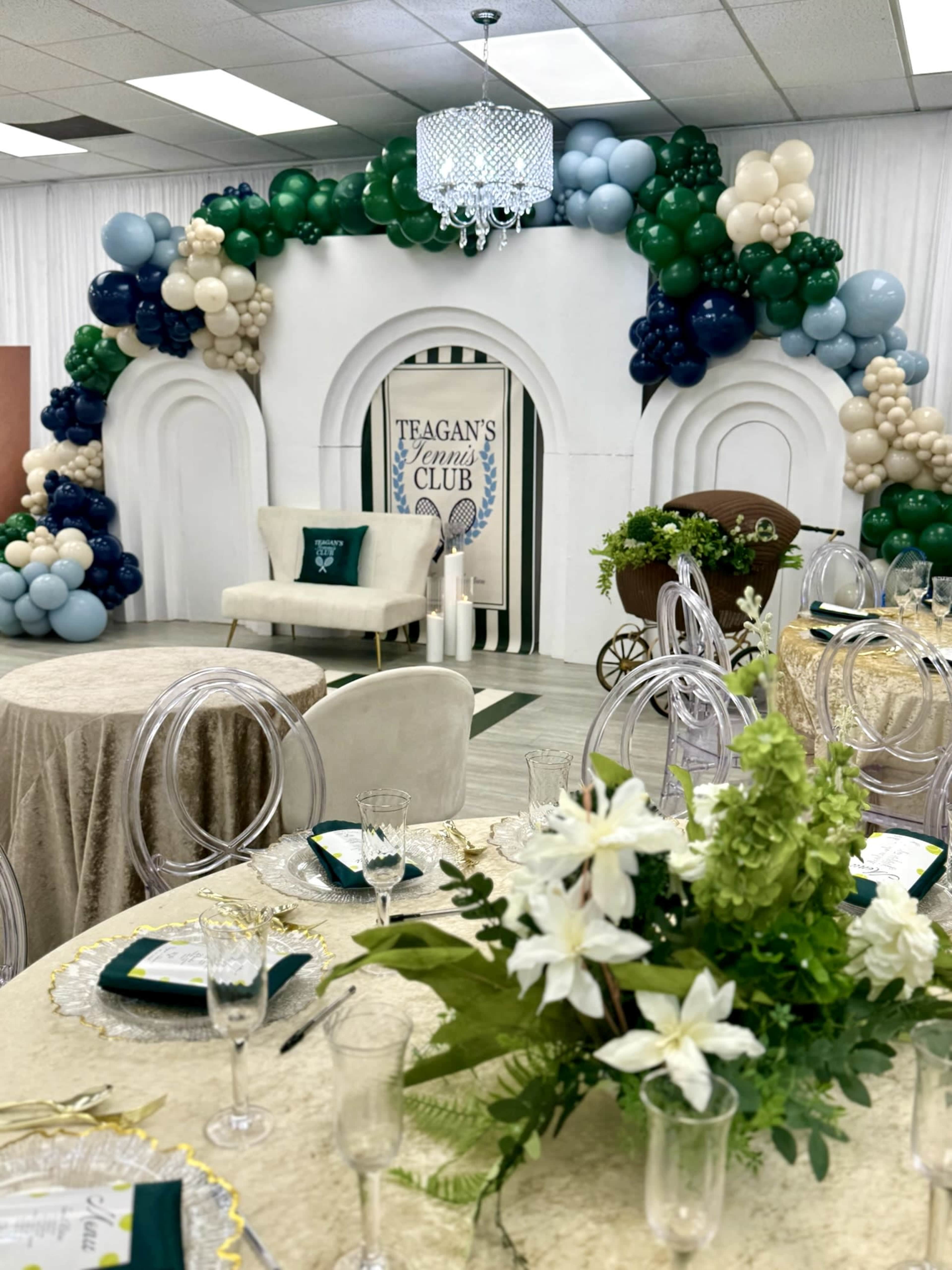The image shows a decorated event space featuring a balloon arch, a sign for "Teagan's Tennis Club," and elegantly arranged table settings with floral centerpieces.