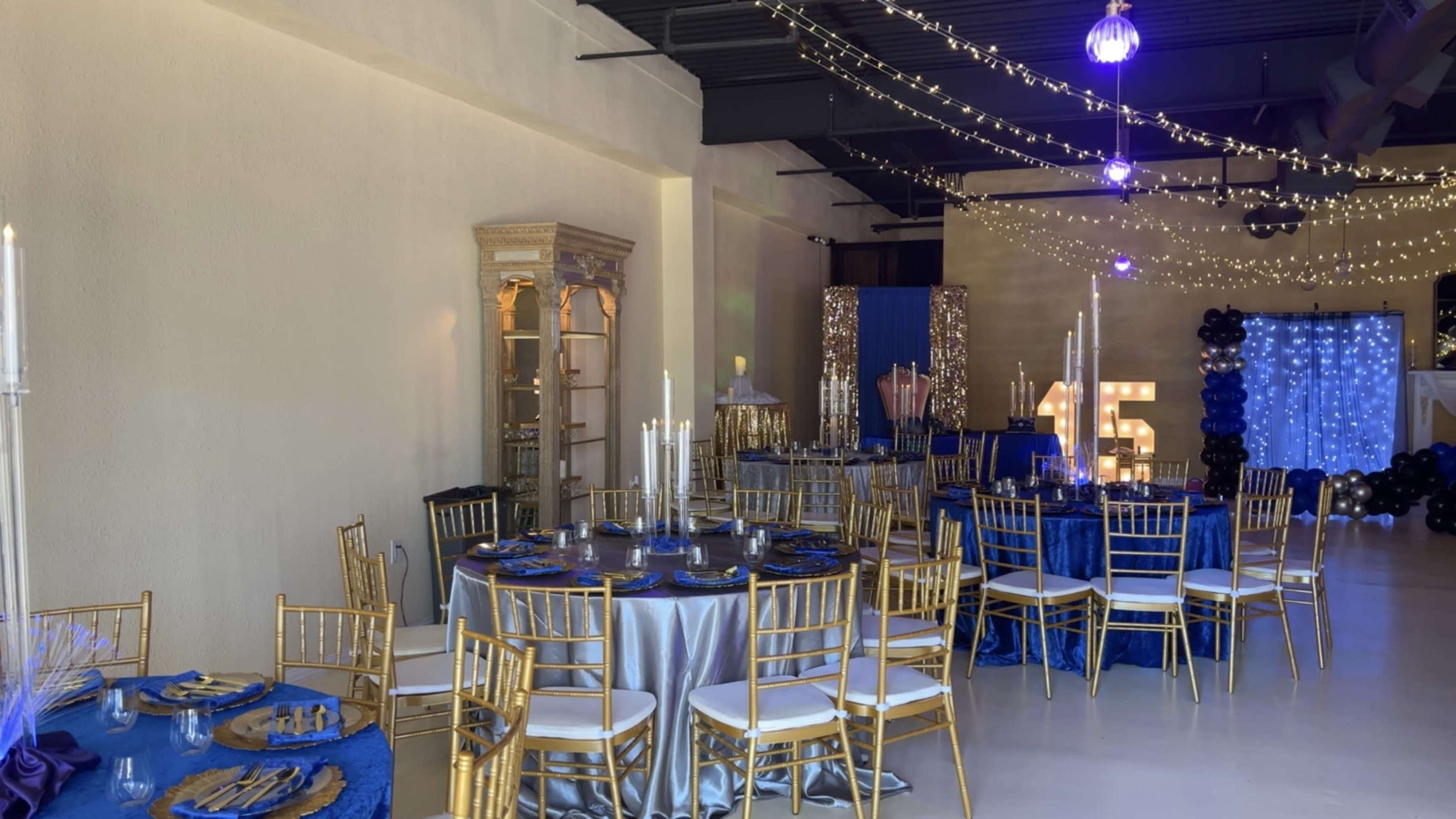 The image shows an elegantly decorated event space featuring round tables with blue and silver tablecloths, golden chairs, and festive lighting embellishments.
