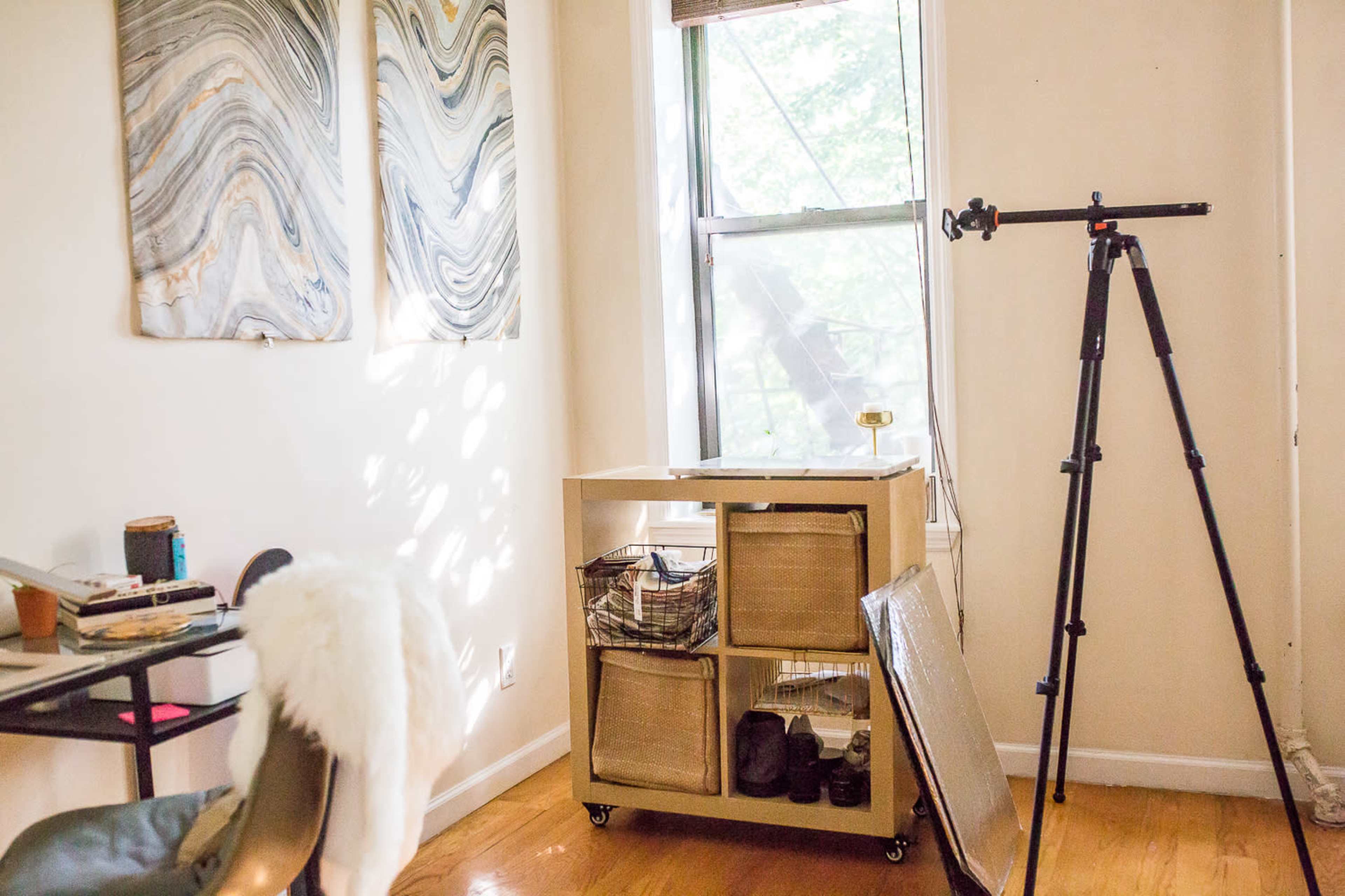 A room features a window with sunlight streaming in, a small table with items on it, a storage cart with baskets, and a tripod standing nearby.