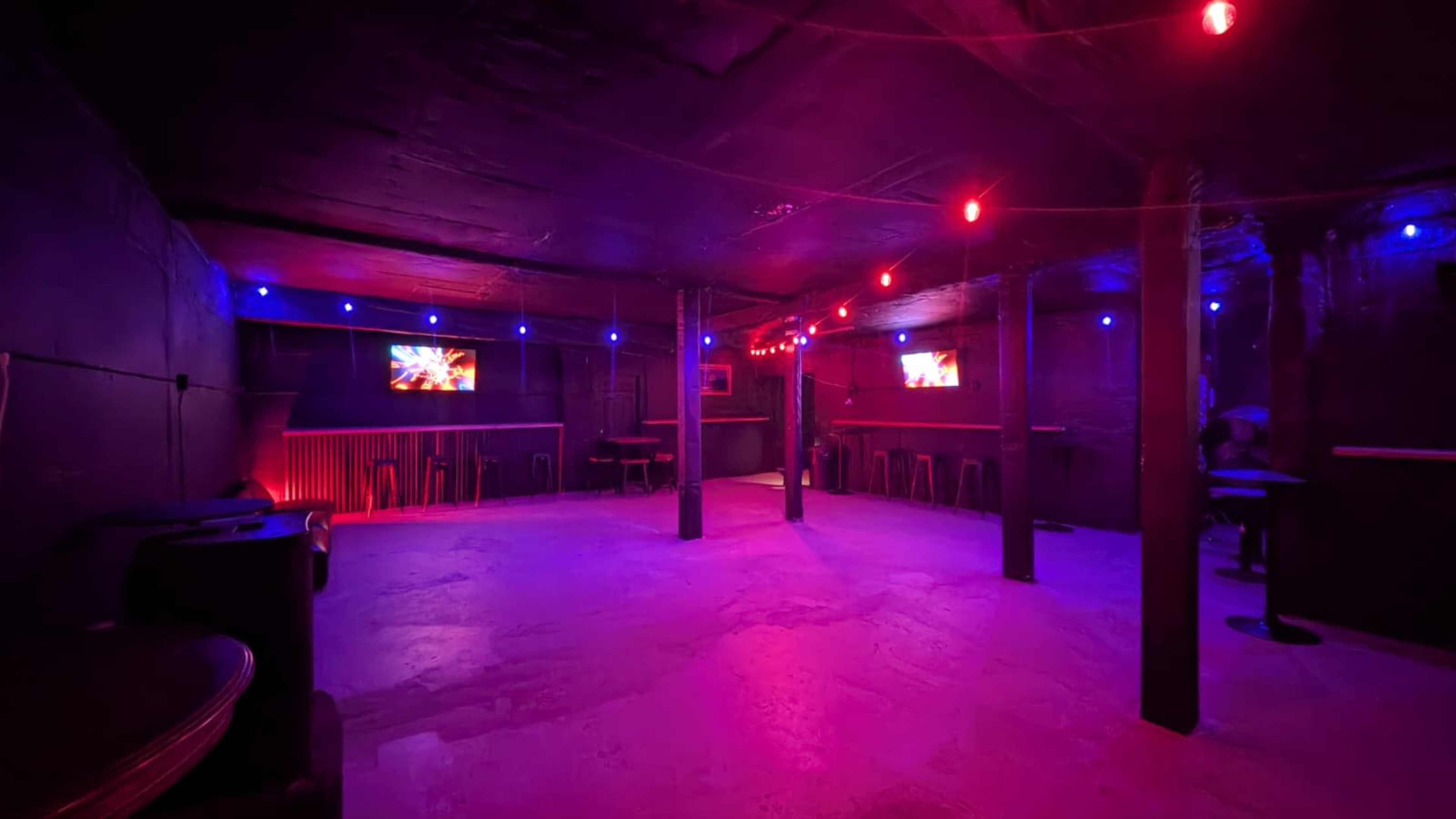 The image shows a dimly lit room with colorful red and blue lights, featuring several tables and screens on the walls.