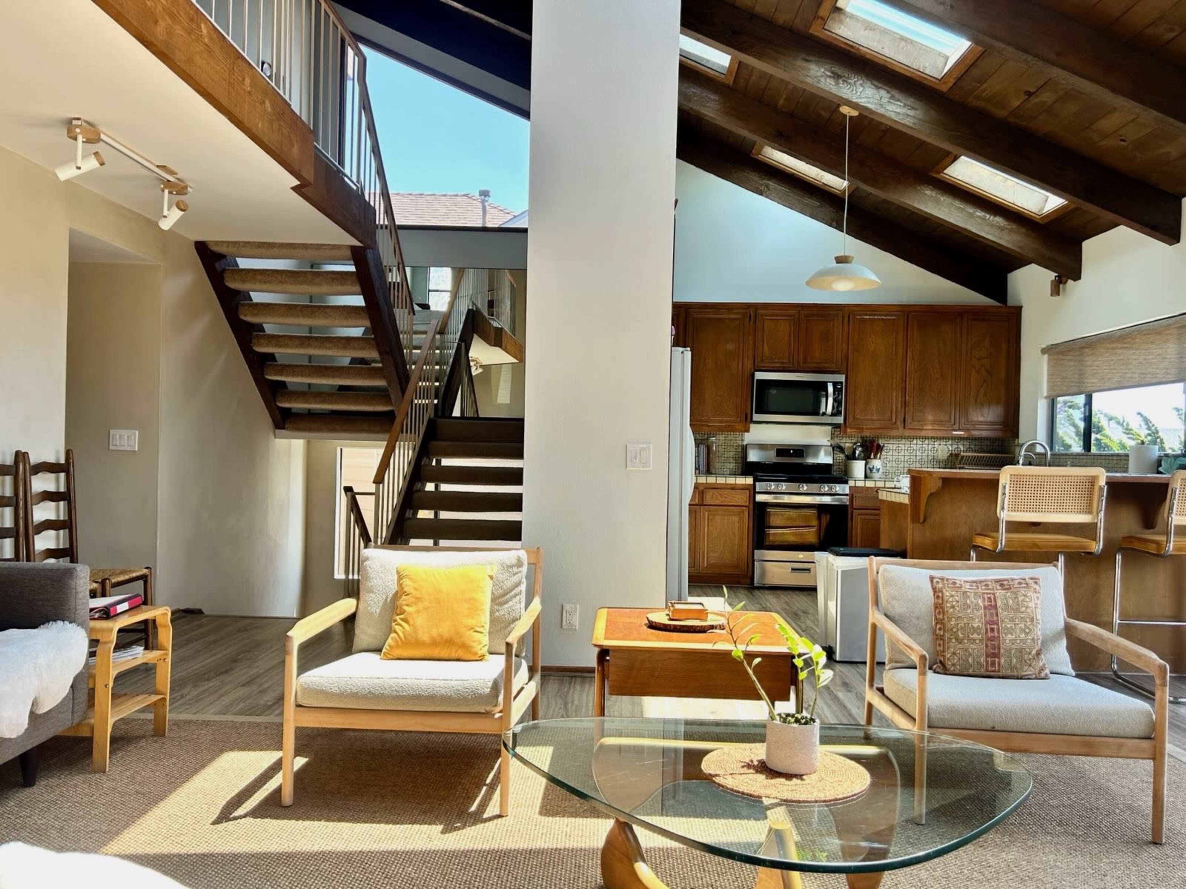 The image shows an open-concept living area with a staircase, modern kitchen, and seating area featuring wooden furniture and large windows.